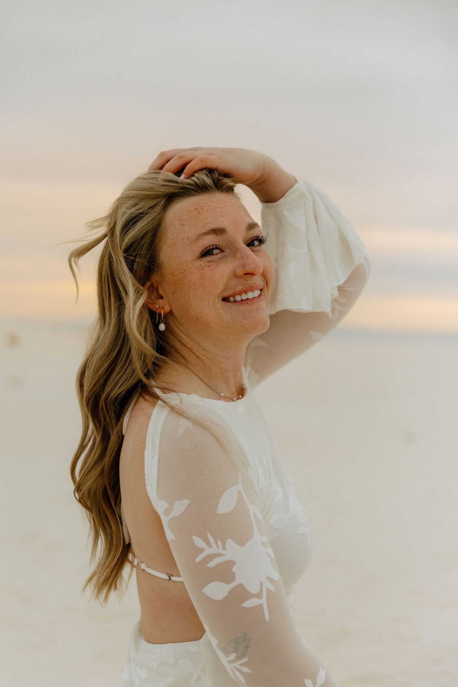 Young woman with freckles in white lace dress laughing while holding hair back on beach at sunset.