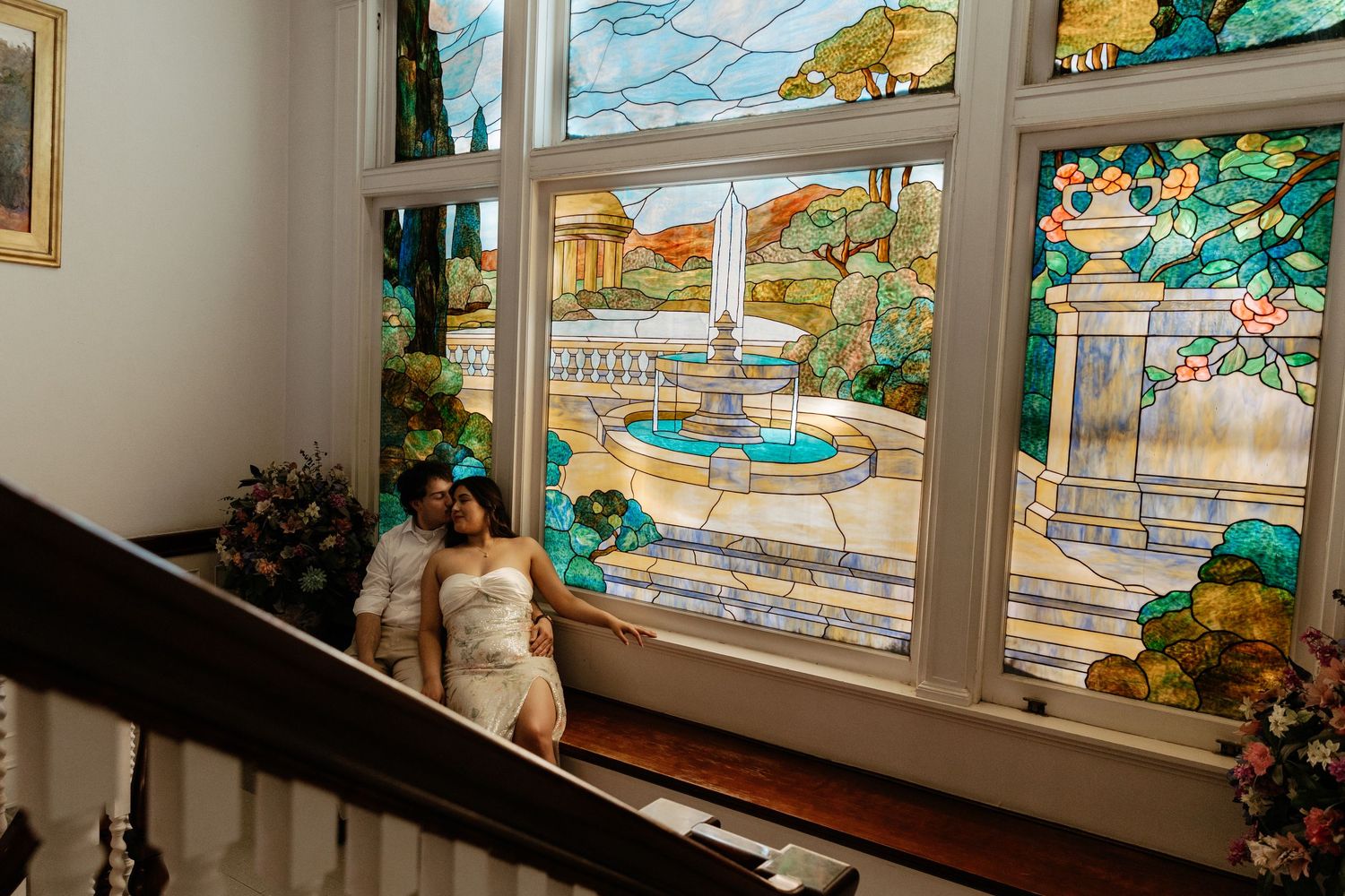 Bride in white gown posing beside ornate stained glass window featuring fountain scene on staircase.