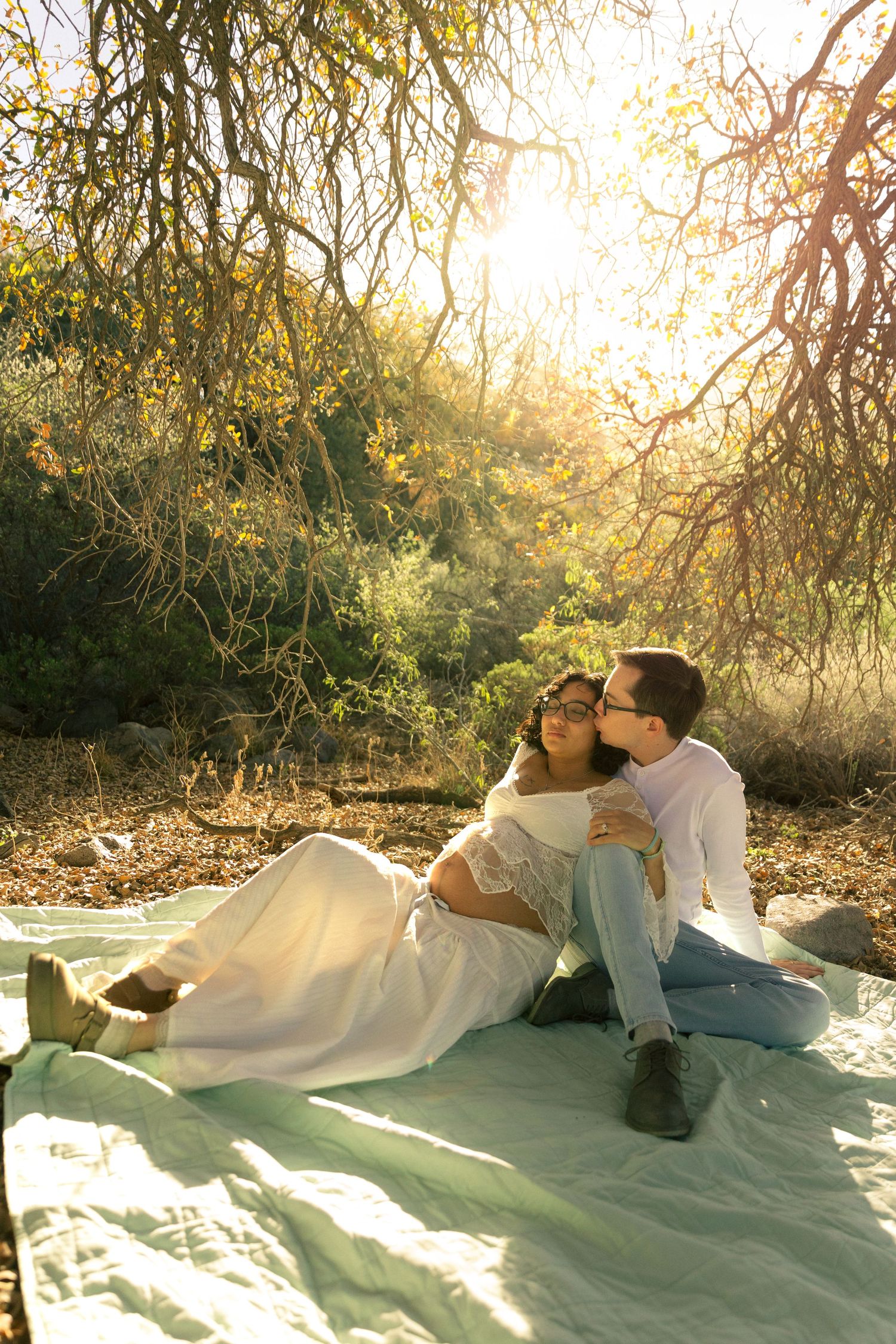 Couple sitting on blanket outdoors, man kissing pregnant woman in white dress, golden sunlight filtering through trees.