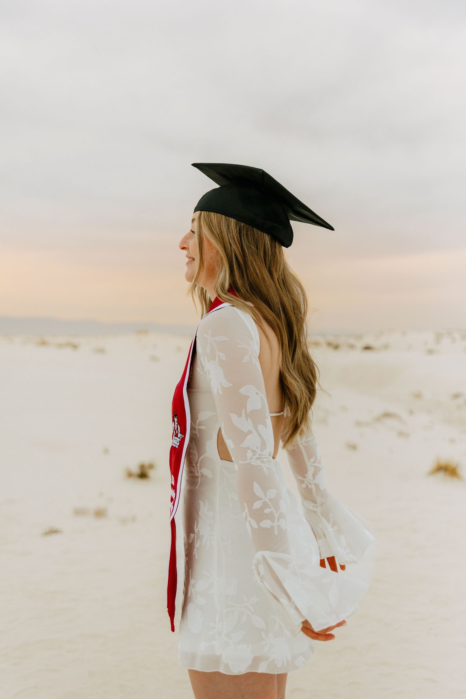 Graduate in white lace dress with red stole and mortarboard cap at White Sands dunes.
