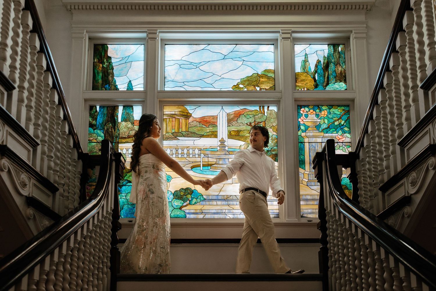 Couple holds hands on a staircase before a stunning stained glass window in an elegant venue.