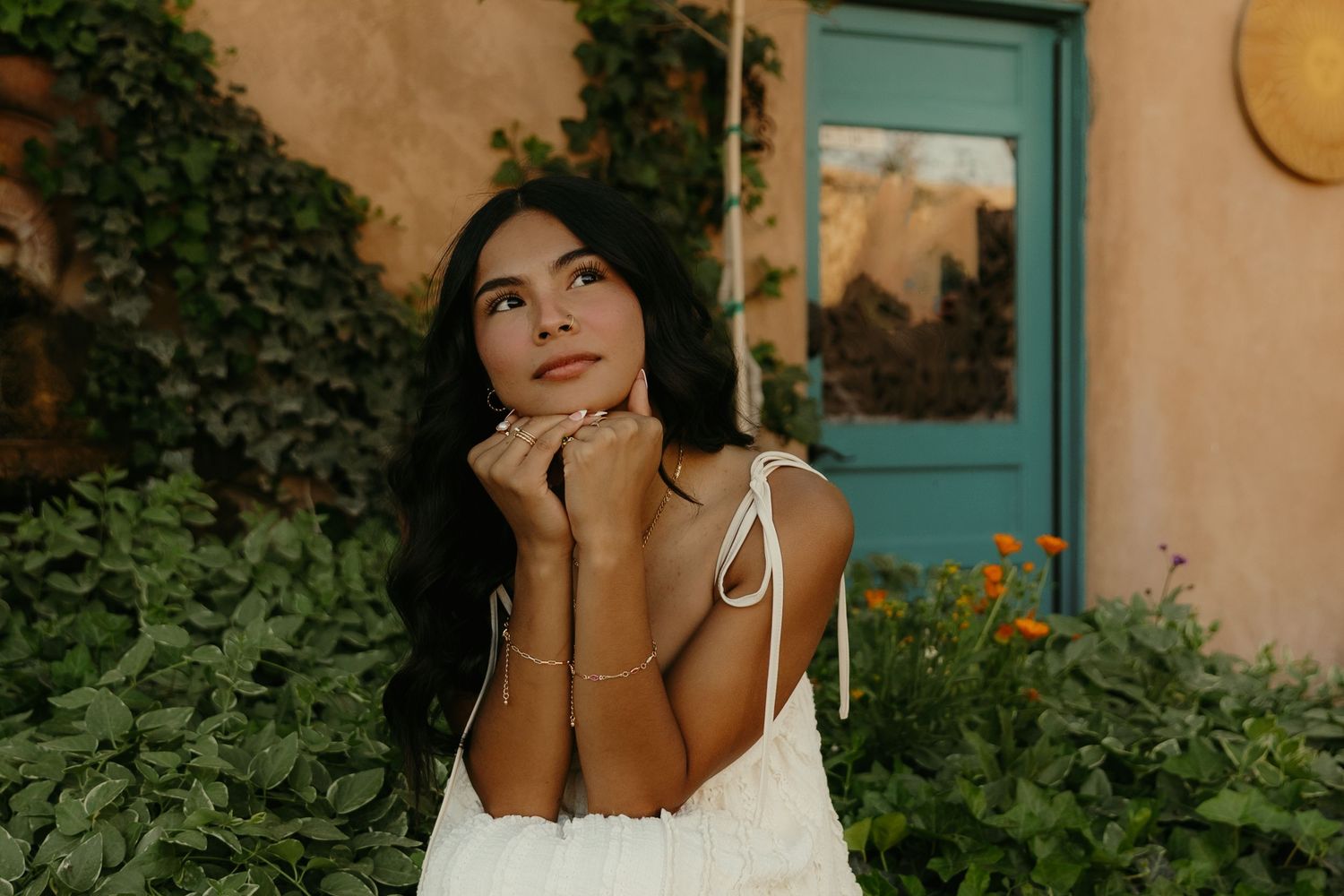 Young woman in white dress poses thoughtfully outdoors near teal door, surrounded by lush green ivy and orange flowers.