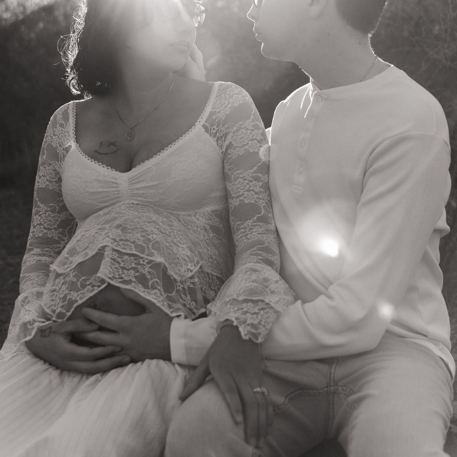 Black and white maternity photo of a couple sitting closely together, the pregnant woman in lace dress, bathed in soft sunlight.