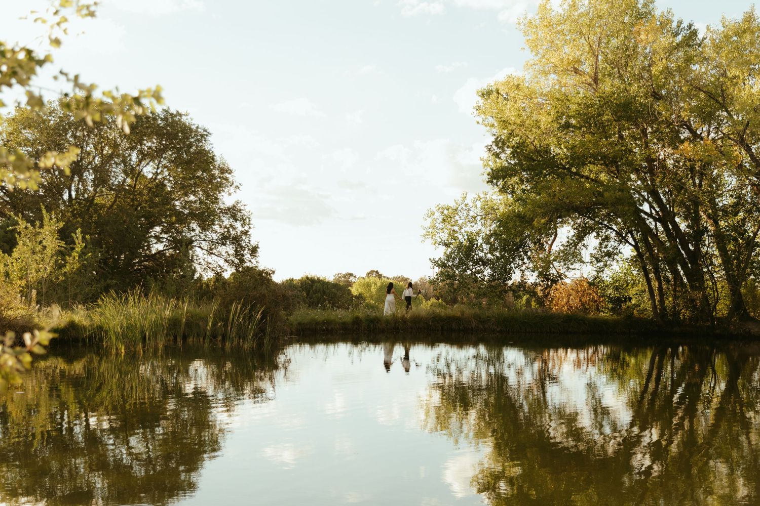 Calm pond reflecting golden autumn trees and a soft blue sky, with two people standing in the distance along the grassy shoreline.
