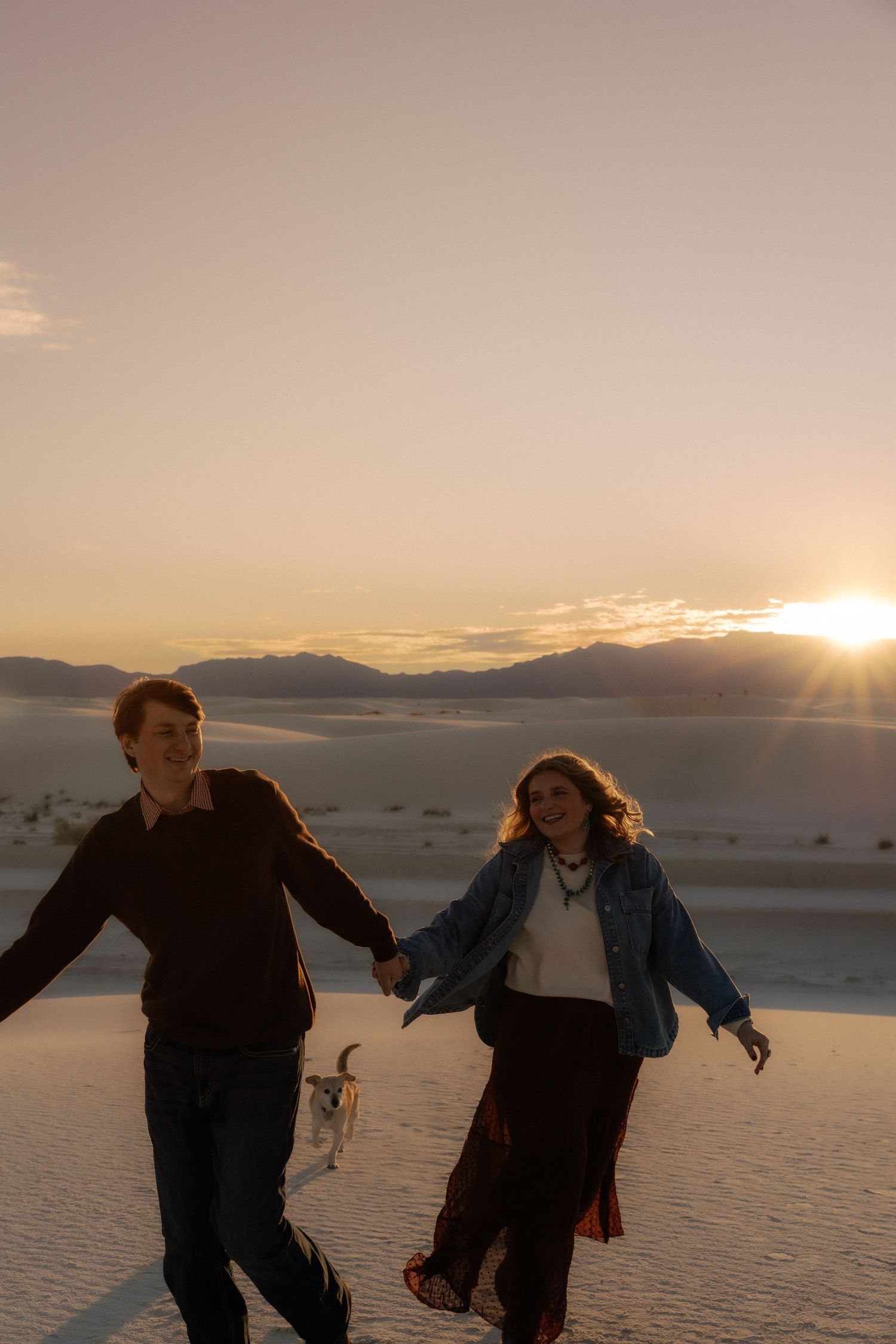 Happy couple holds hands walking on white sand dunes with dog at sunset.