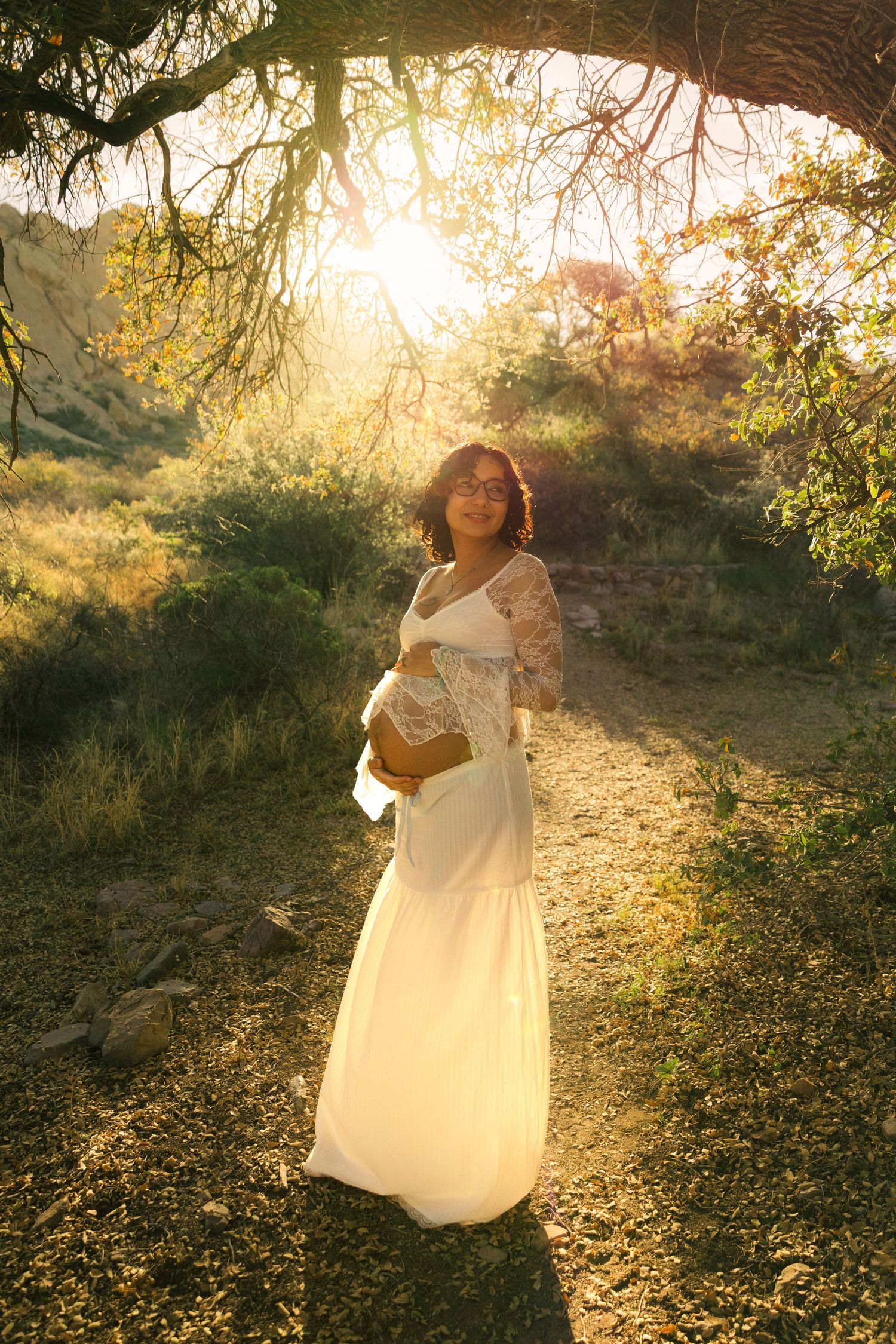 Pregnant woman in white lace gown standing in sunlit nature path, cradling baby bump during golden hour maternity shoot.