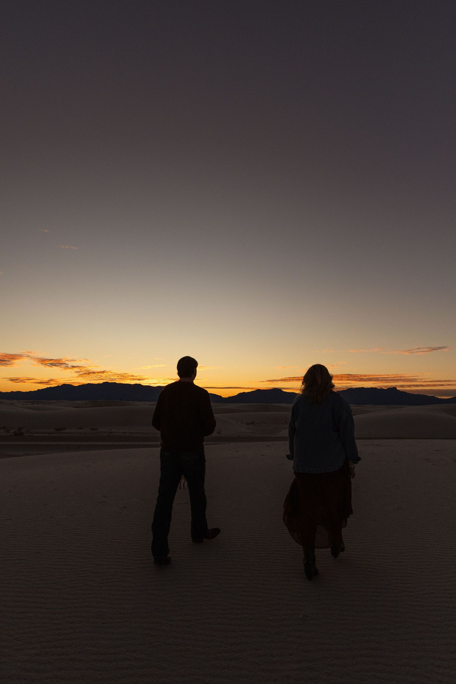 Silhouette of couple standing on sand dunes watching a vibrant orange and gold desert sunset together.