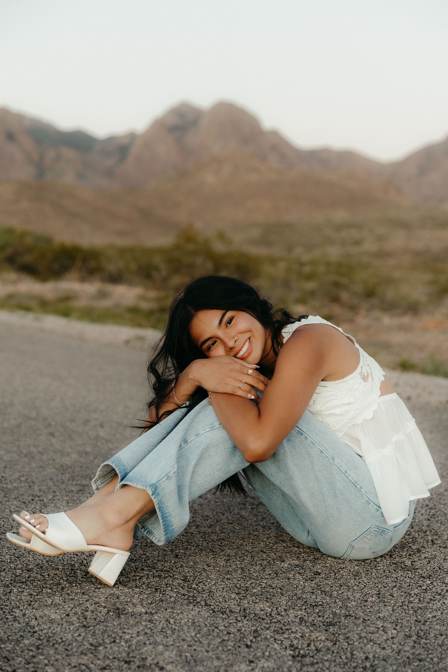 Smiling woman in white top and jeans sitting on road with scenic mountain landscape behind her.