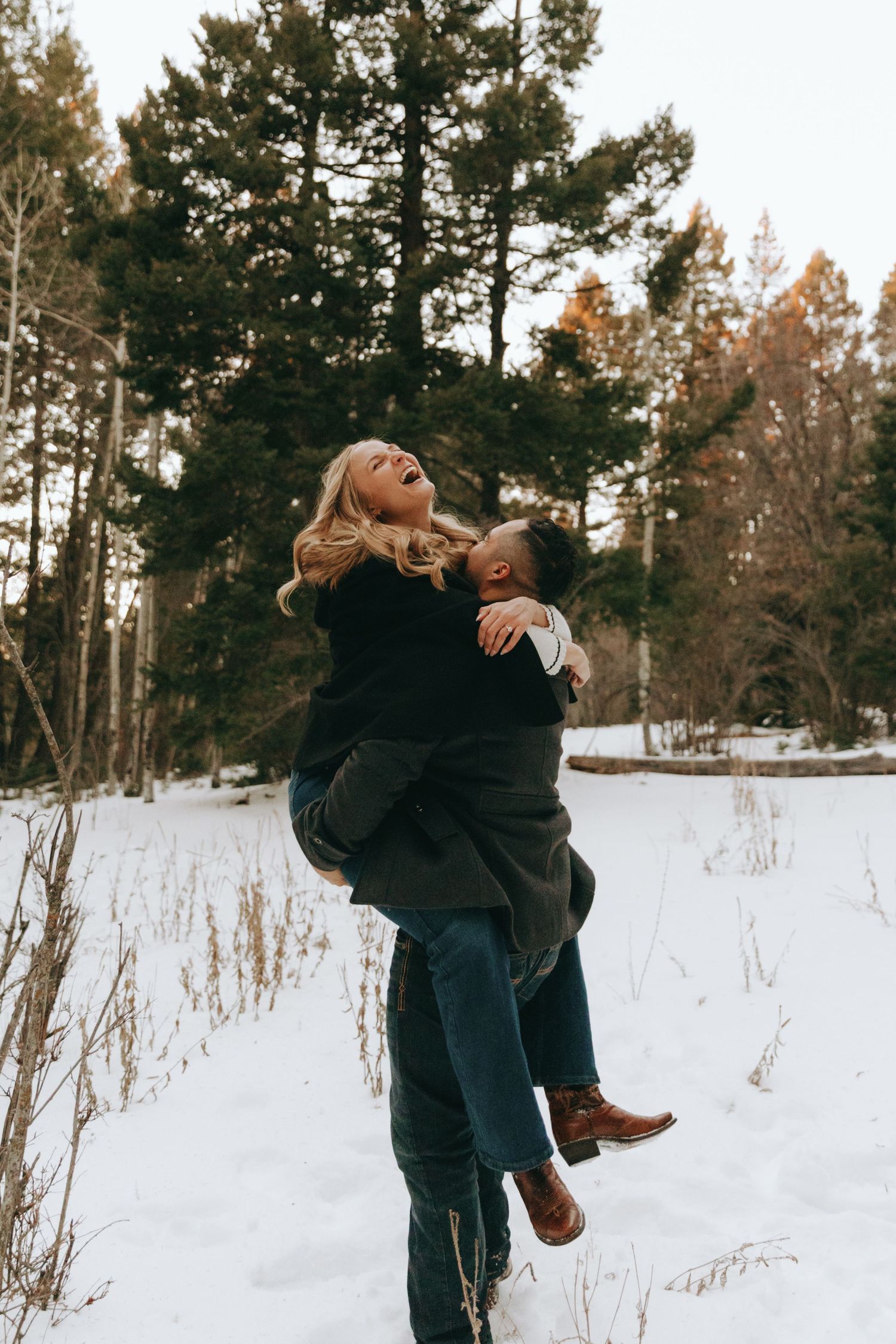 Couple laughing joyfully in snowy winter forest, man lifting woman in black coat, pine trees background.