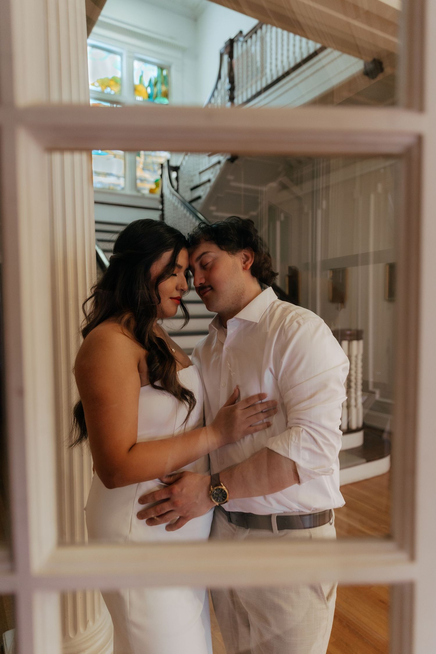 Couple sharing an intimate moment framed by a white window, dressed in white wedding attire inside an elegant mansion.
