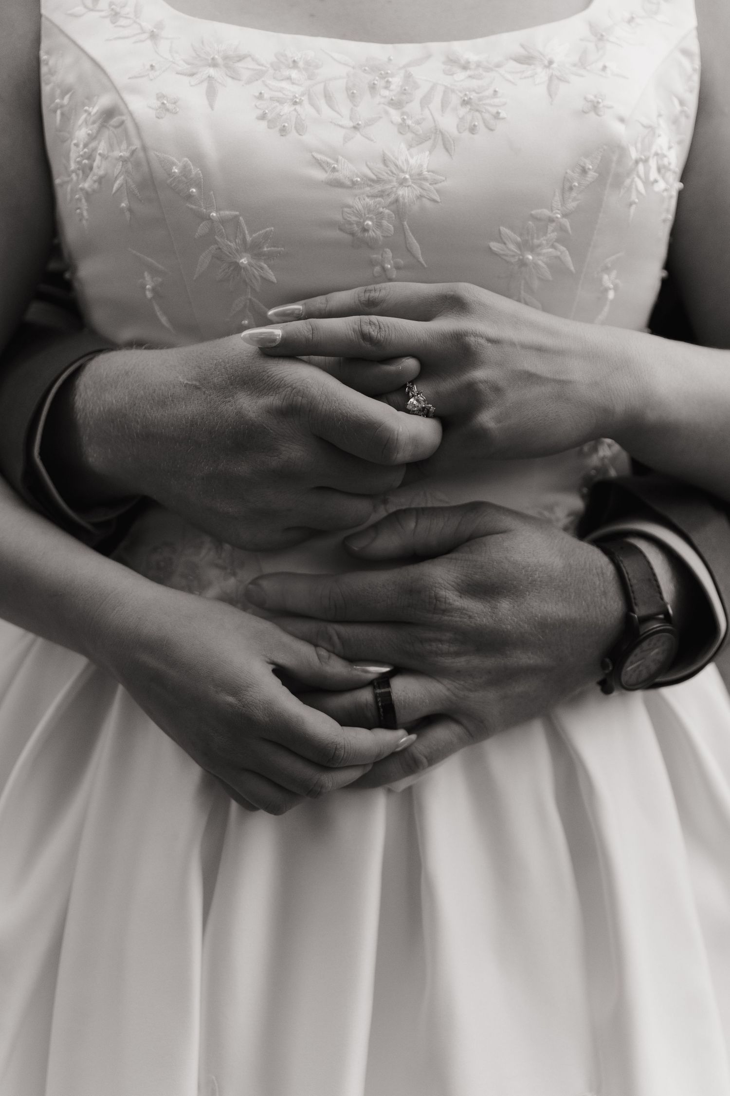 Black and white photo of bride and groom holding hands, showing wedding rings and a watch, against white bridal gown.