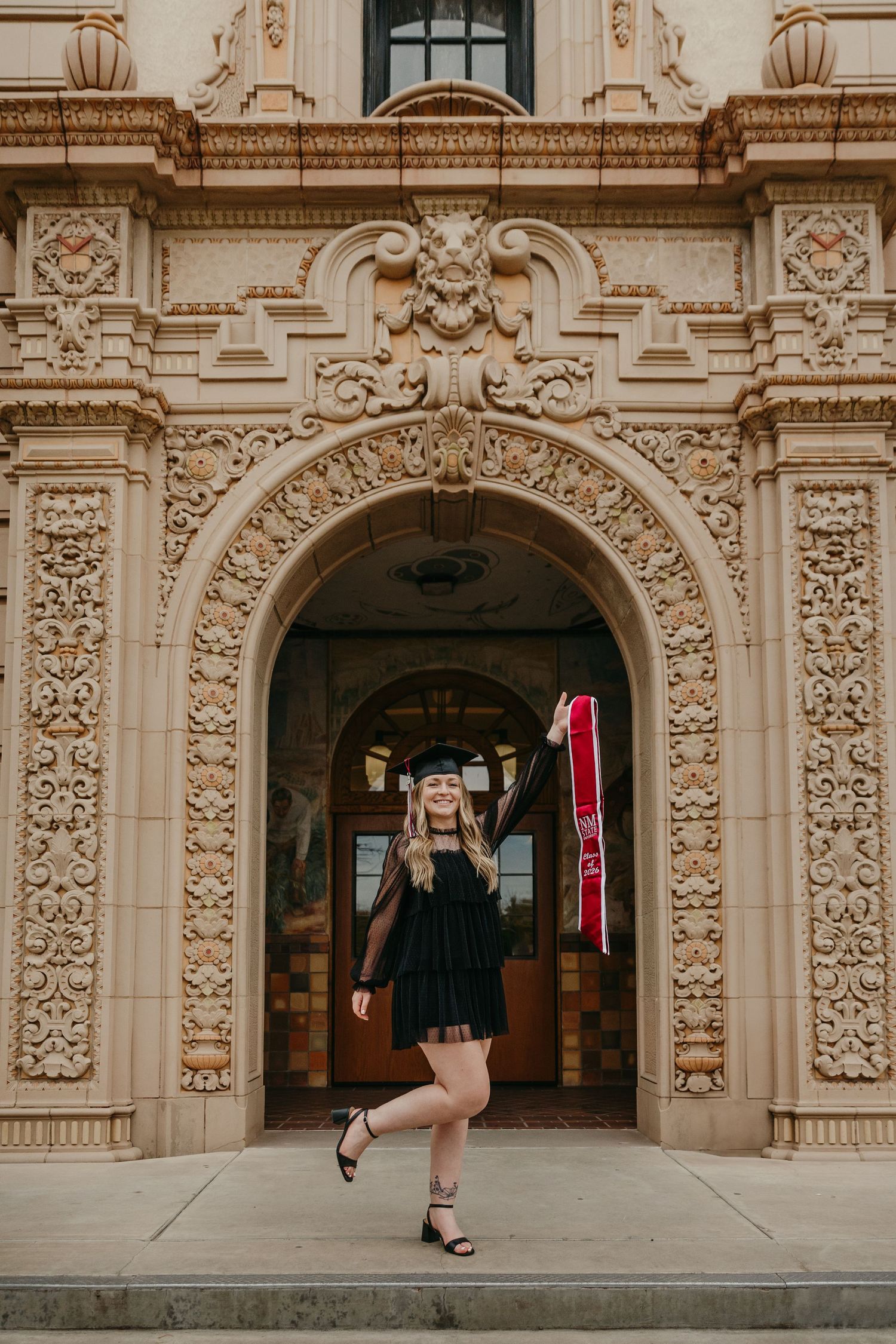 Graduate in black dress with red stole and cap posing at ornate building entrance.