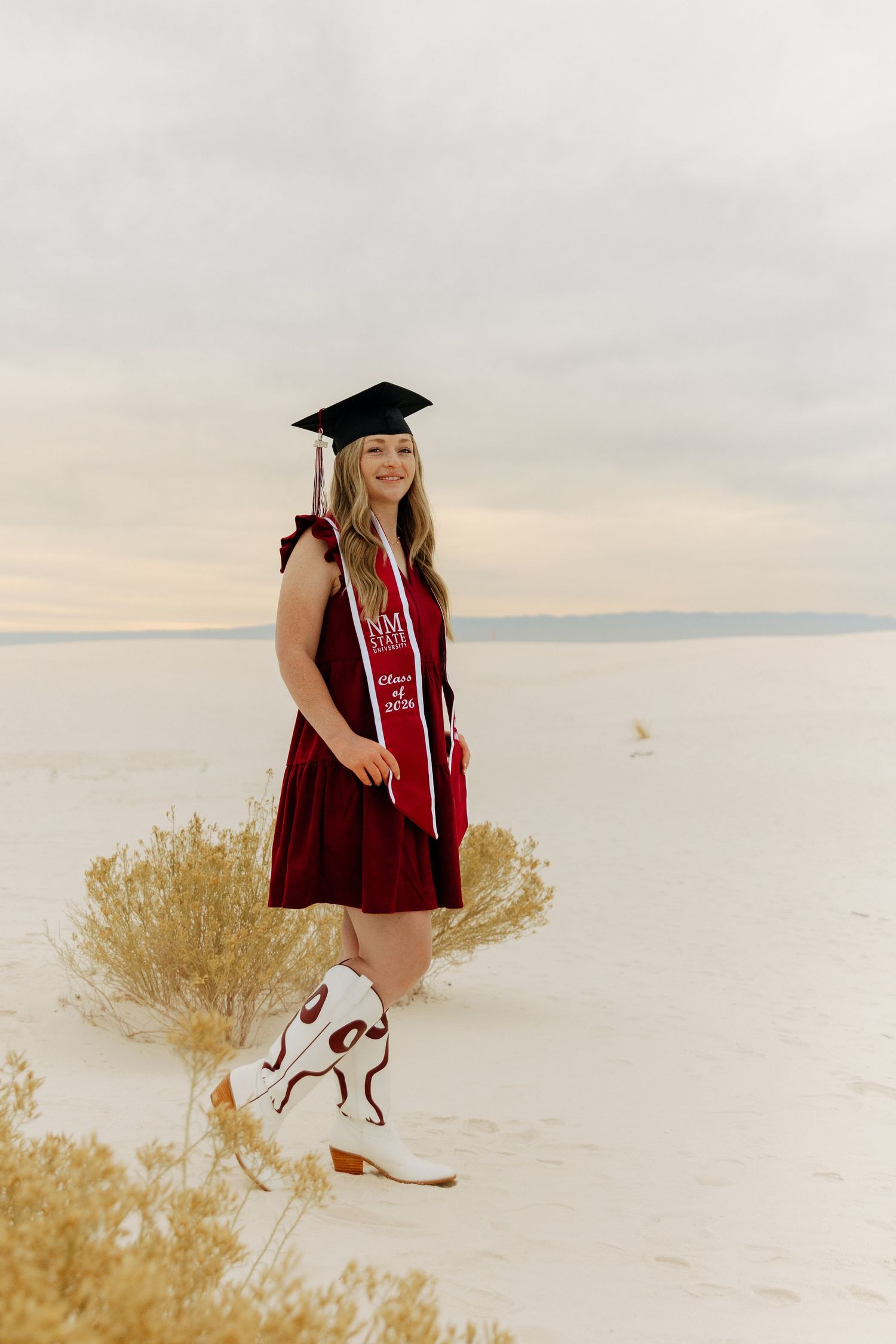 Graduate in maroon dress, cap, and white cowboy boots at White Sands National Park.