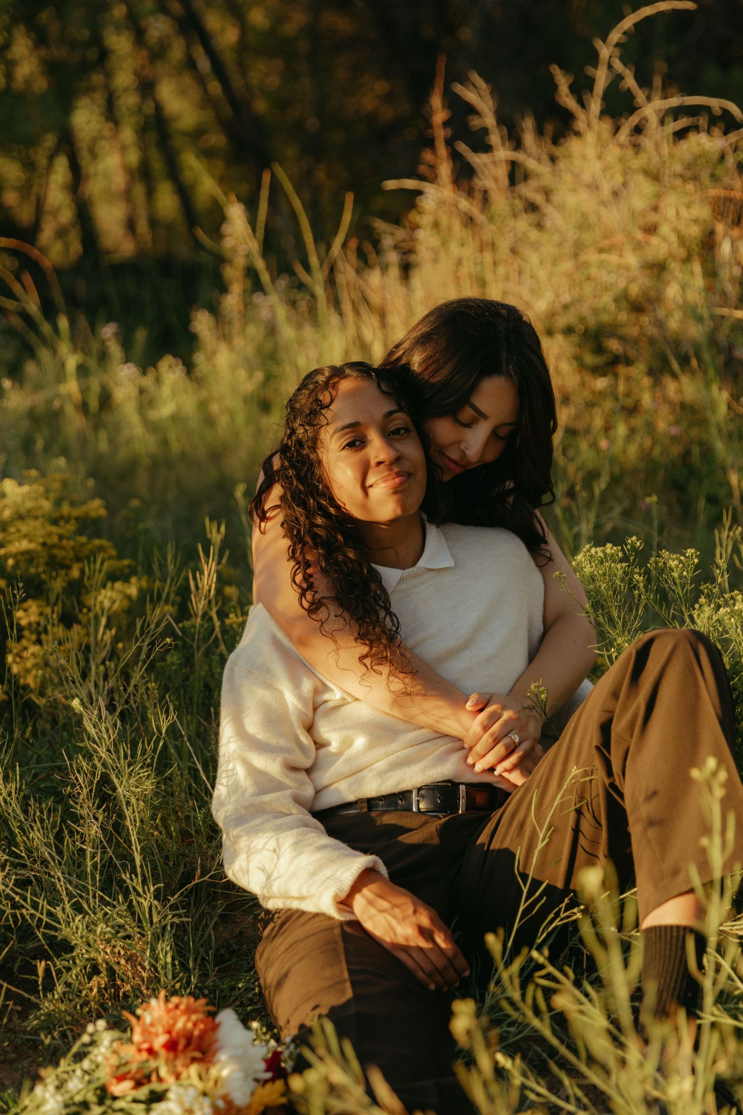 Two women embrace affectionately while seated in a golden-lit meadow of tall grass, with a colorful bouquet of flowers resting nearby.