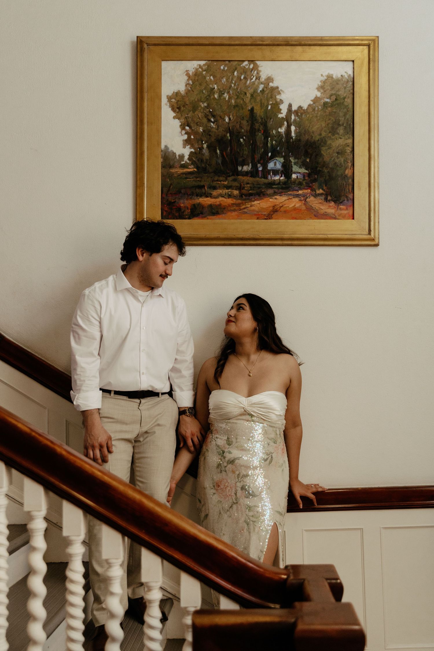 Couple in wedding attire sharing a loving gaze on a classic staircase beneath a landscape painting.
