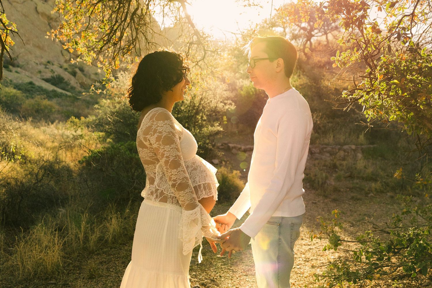 Pregnant couple holding hands in golden sunlight among autumn trees, woman in white lace maternity gown.