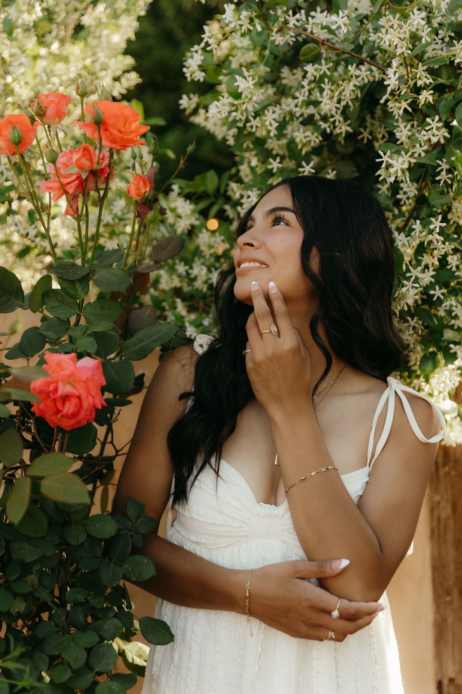 Young woman in white dress smiling among orange roses and white flowering vines in garden.