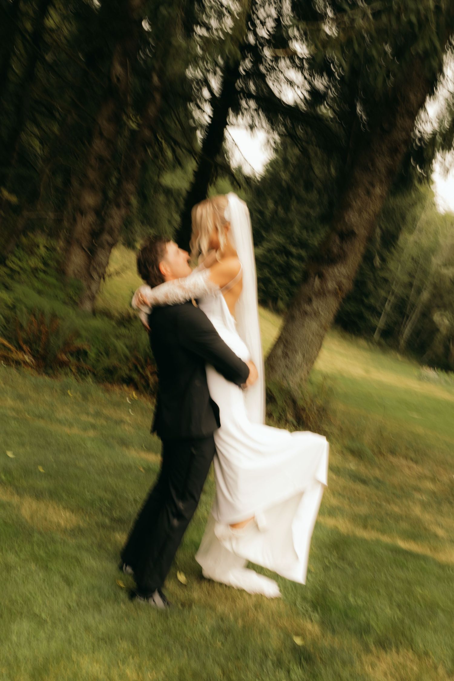 Blurry motion shot of a bride and groom embracing and kissing outdoors on green grass under large trees.