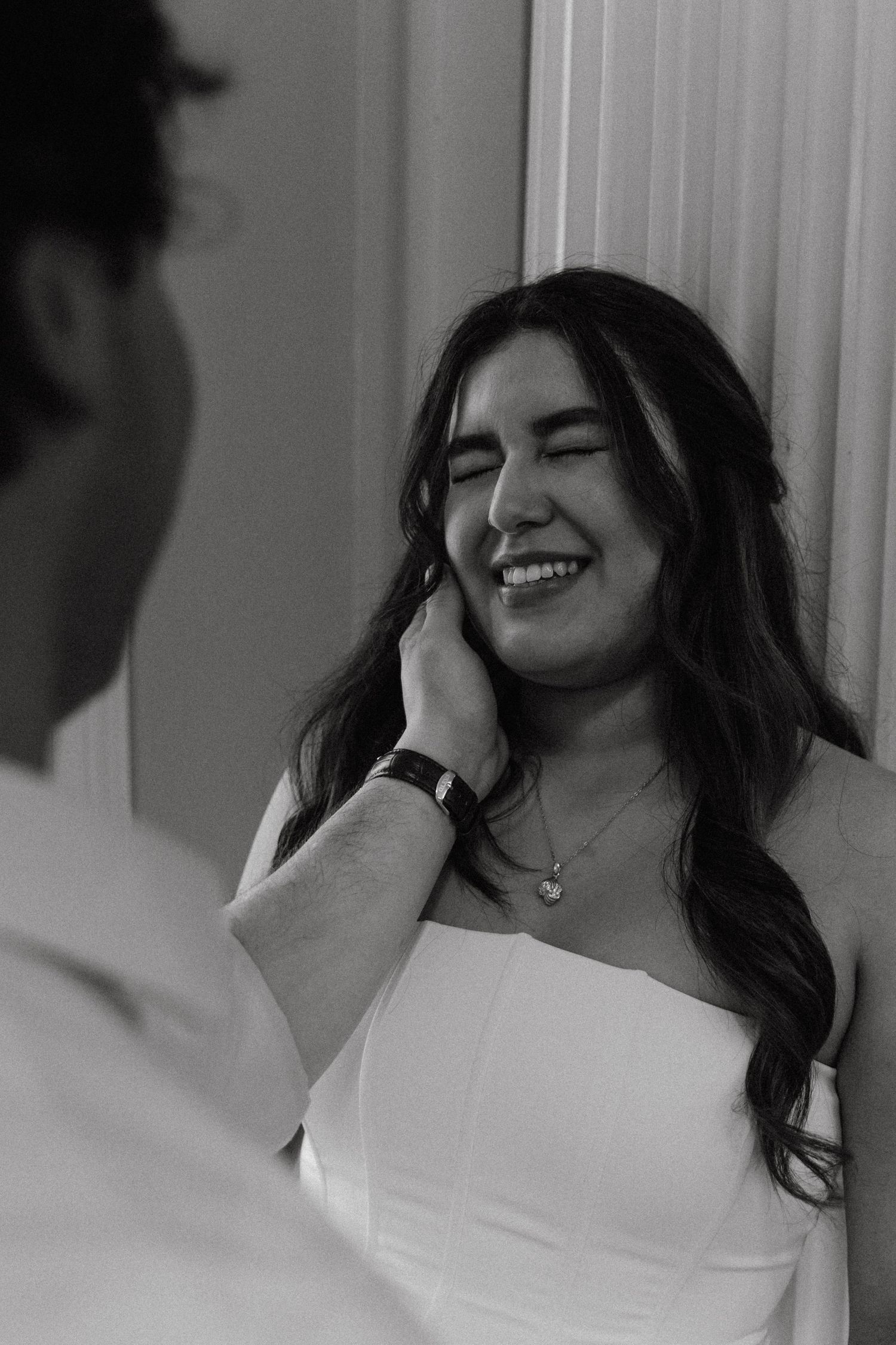 Black and white photo of a groom tenderly touching his laughing brides face indoors.