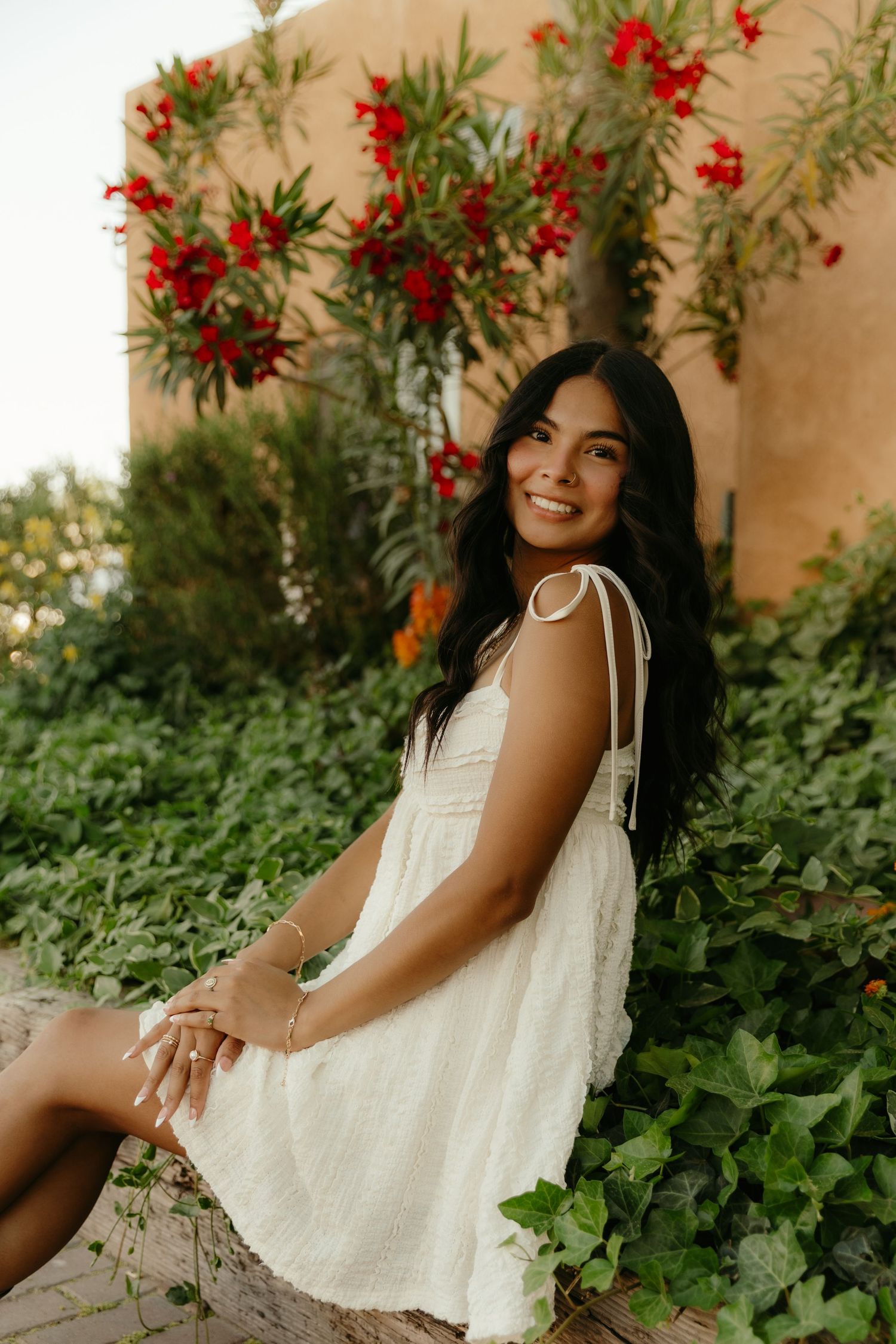 Young woman in white lace dress sitting among green ivy and red flowers, smiling outdoors.