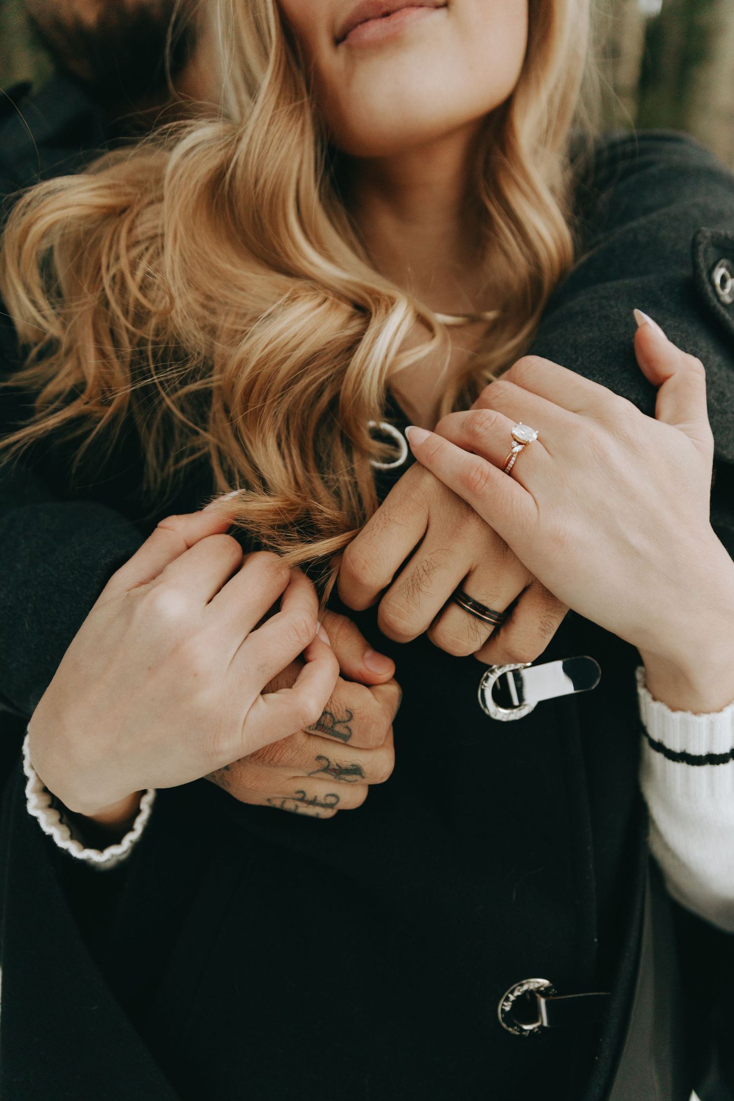Couple holding hands showing engagement ring and wedding band, woman with blonde hair wearing black coat and white sweater.