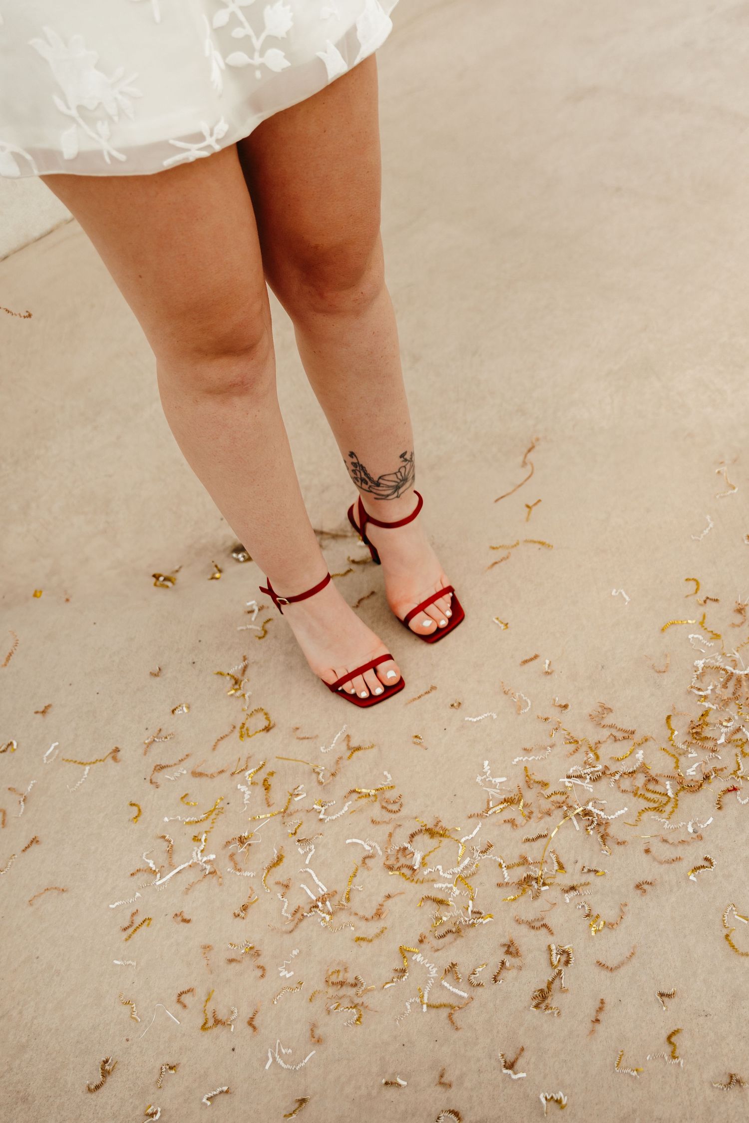 Close-up of womans legs in red strappy heels with an ankle tattoo standing on ground covered in gold confetti.