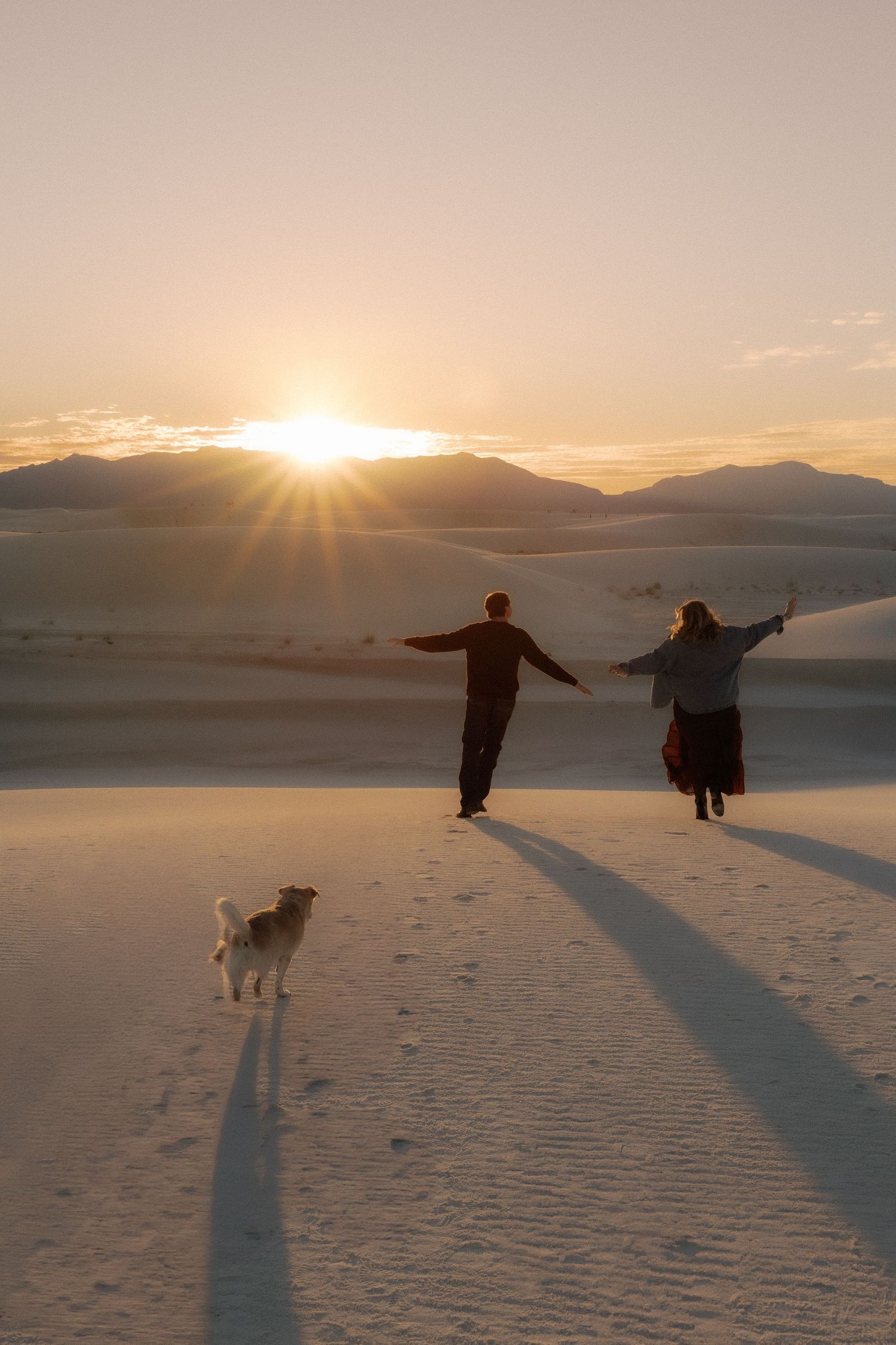 Couple and dog running toward sunset over white sand dunes.