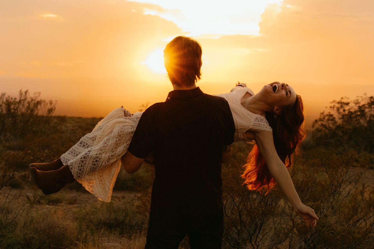 A man lifts a woman in a white dress against a golden sunset sky in an open field.
