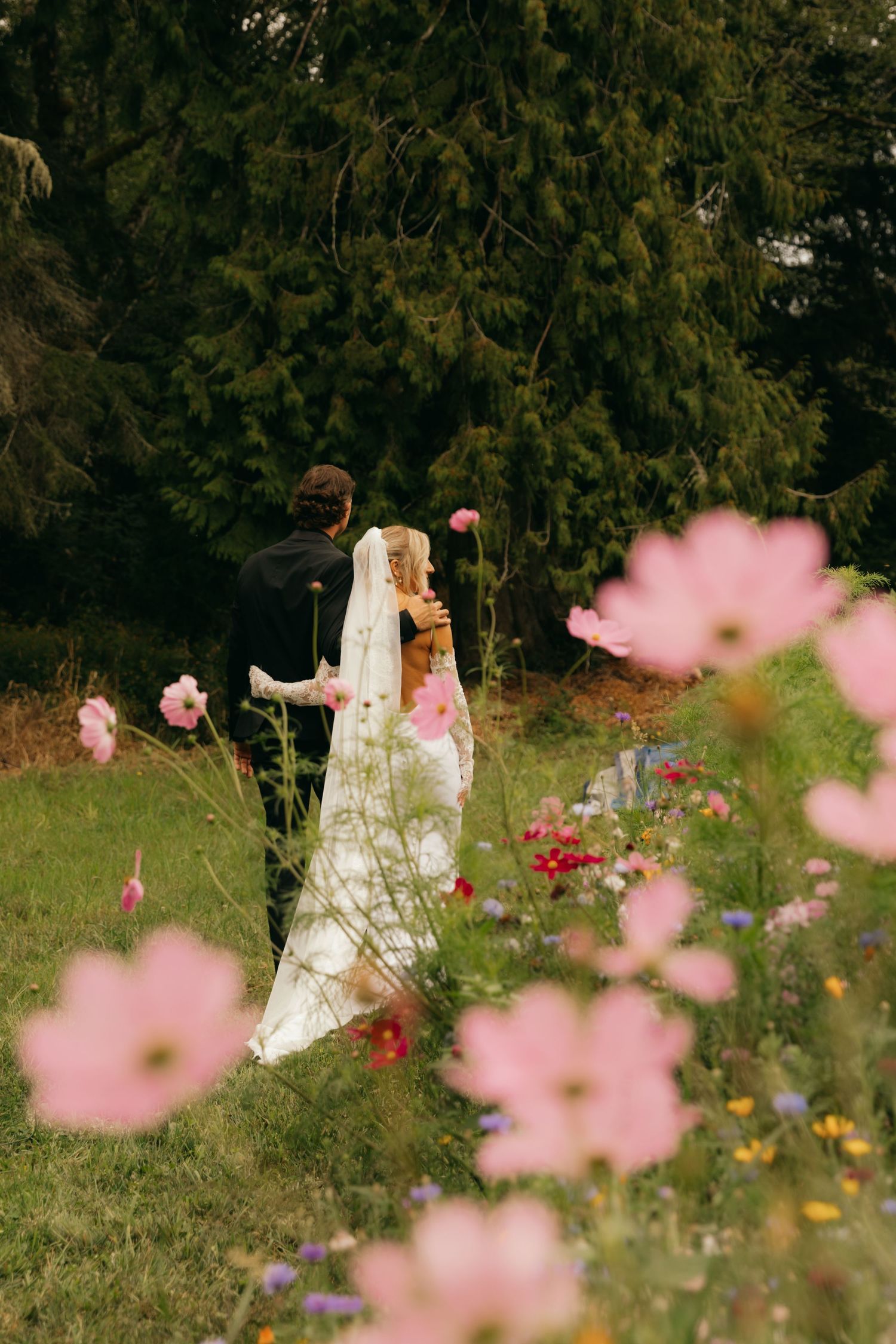Bride and groom walking through a lush garden with pink wildflowers in the foreground on their wedding day.