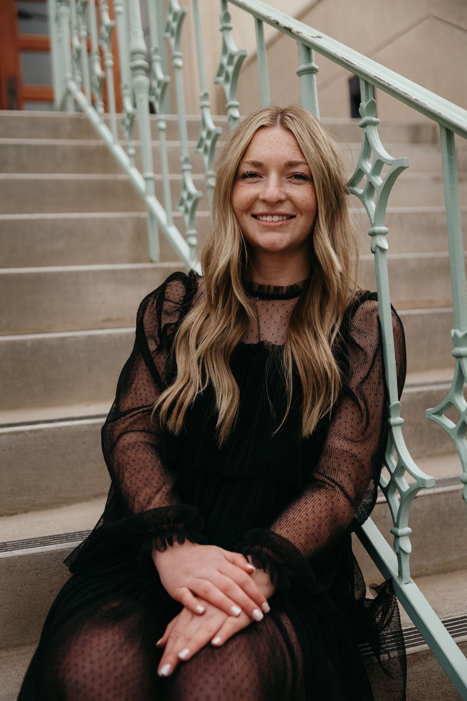 Smiling blonde woman in black lace dress sitting on ornate staircase steps.
