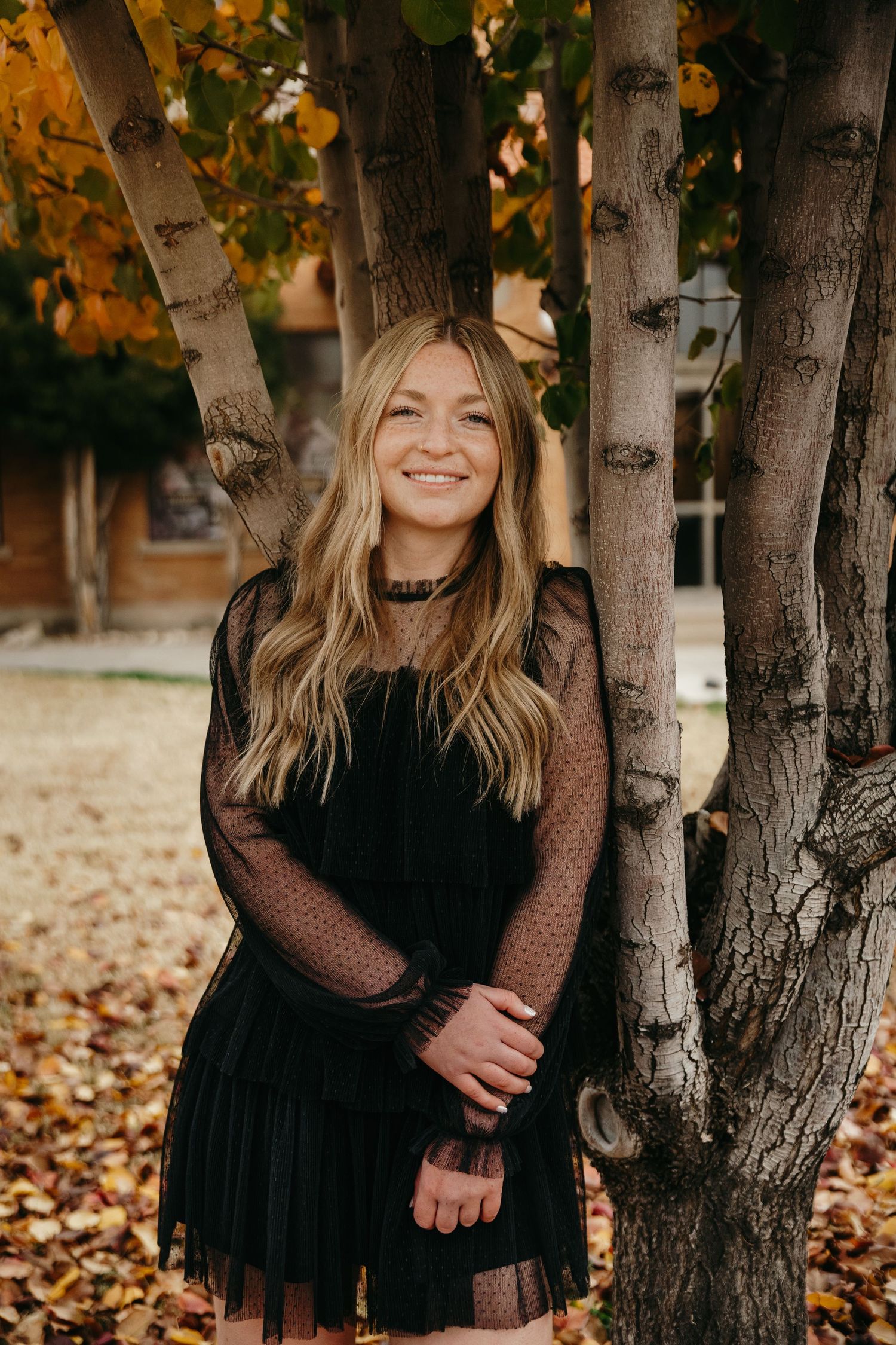 Blonde woman in black sheer dress smiling among birch trees with fall foliage.
