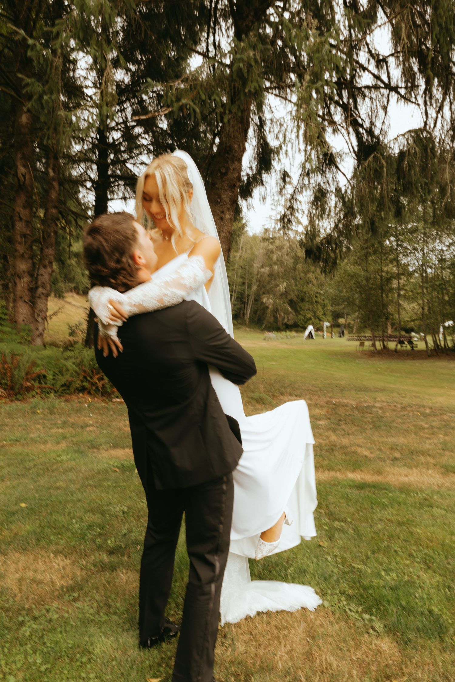 Groom lifting bride in white gown and veil outdoors under large evergreen tree on lush green lawn.
