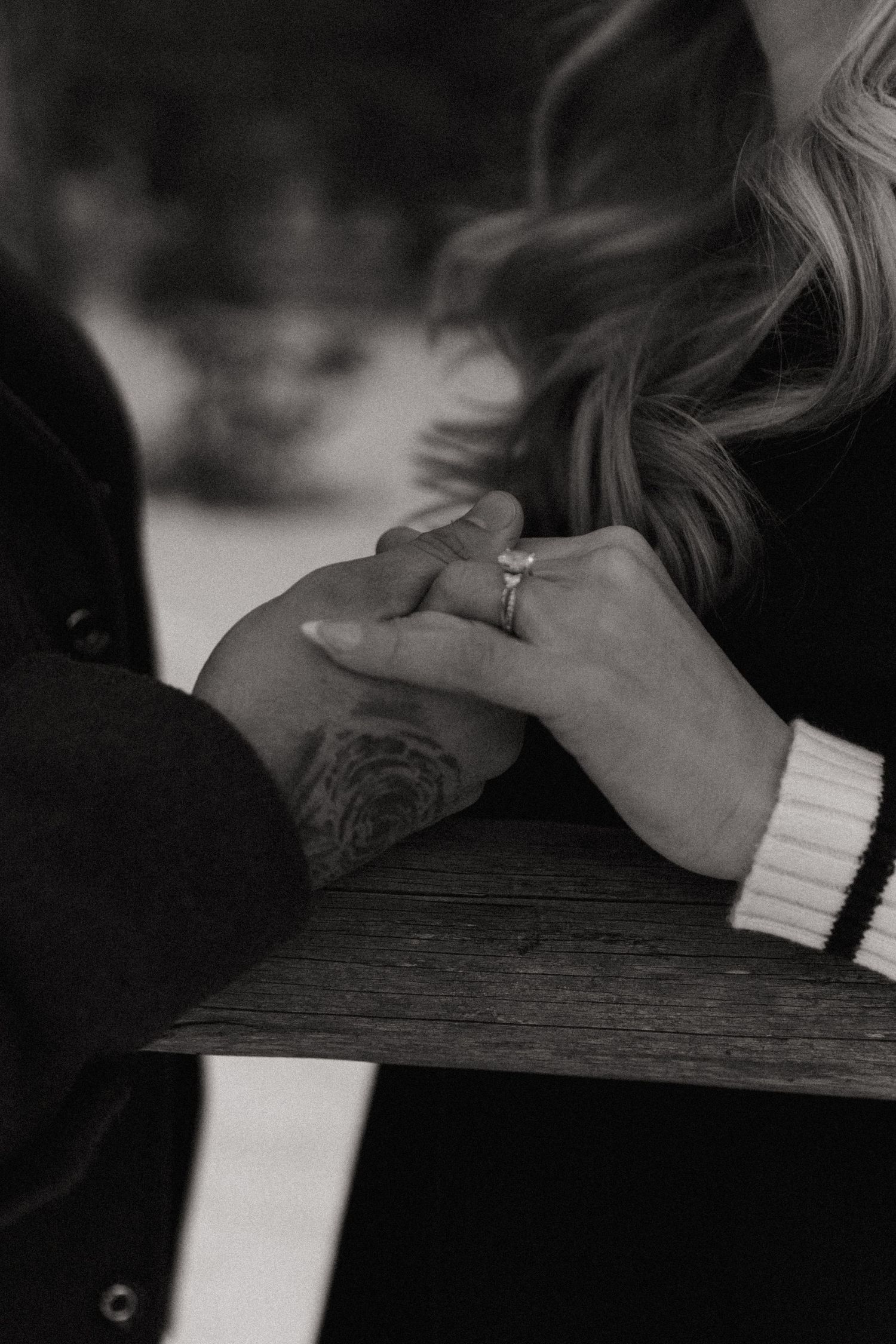 Black and white photo of couple holding hands, showing engagement ring and tattoo on bench.