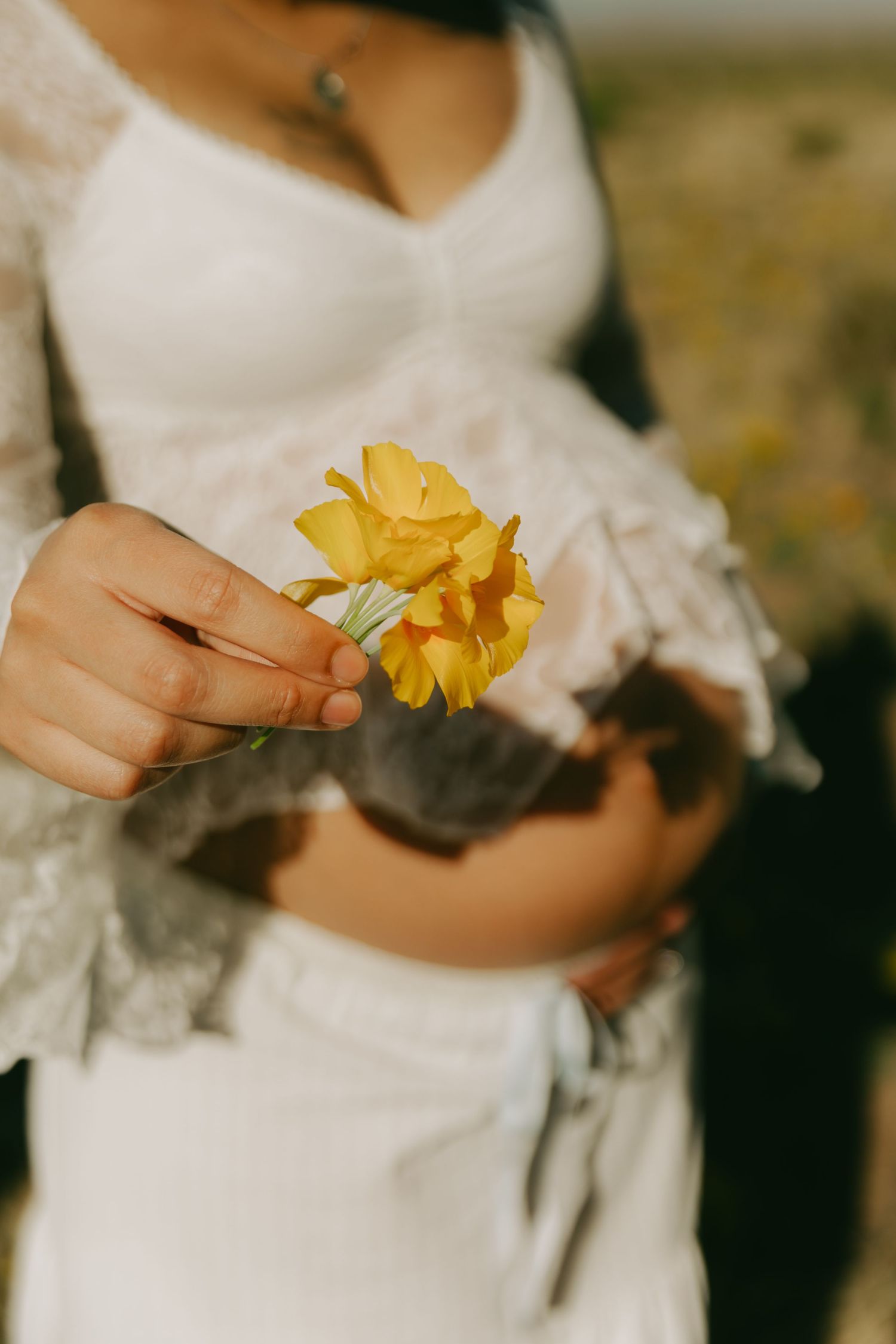 Pregnant woman in white lace crop top holding a yellow daffodil flower outdoors in soft sunlight.
