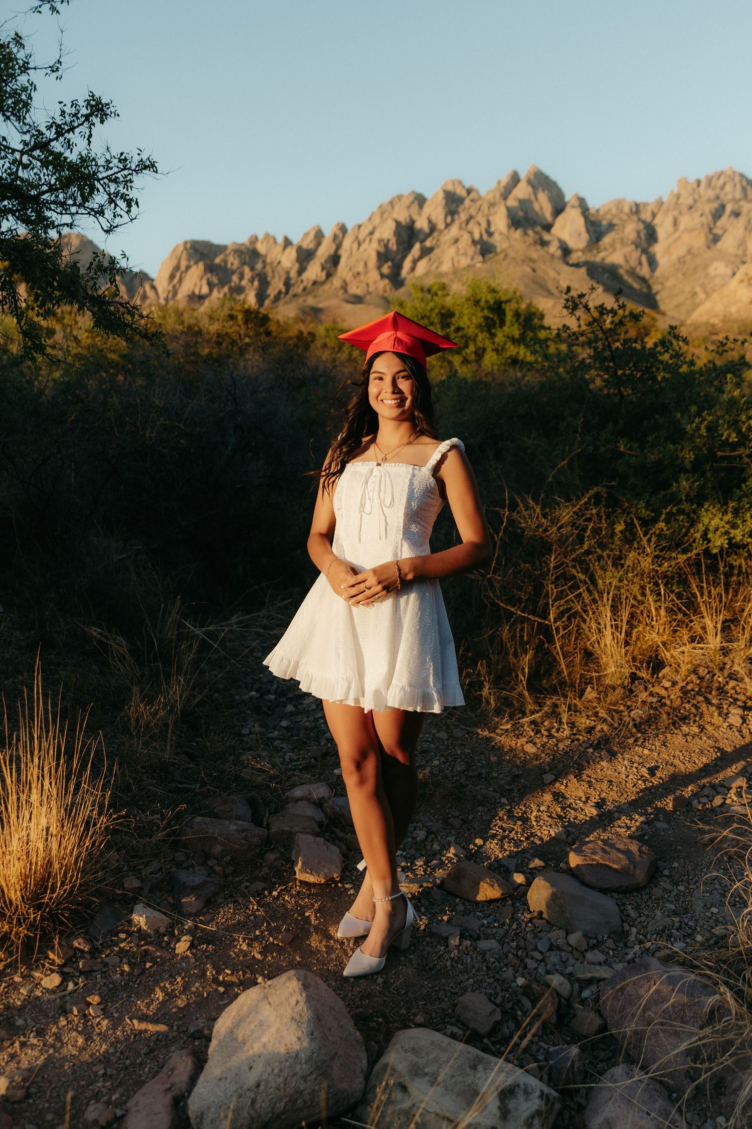 Young woman in white dress and red graduation cap smiling outdoors with rocky mountain landscape at golden hour.
