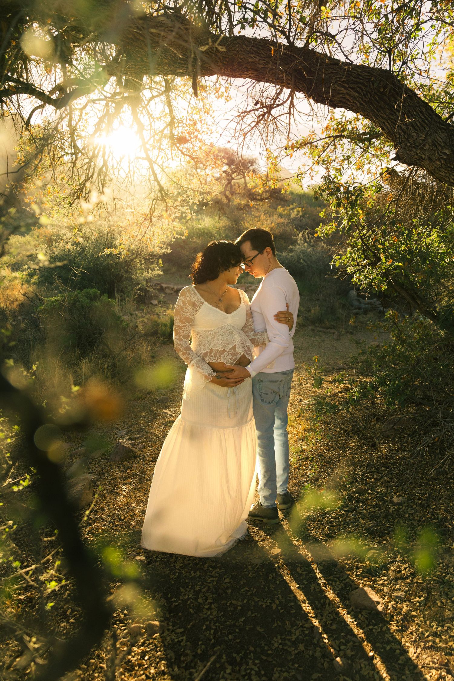 Couple embracing in golden sunset light among lush green trees, woman in white lace gown, man in jeans and white shirt.