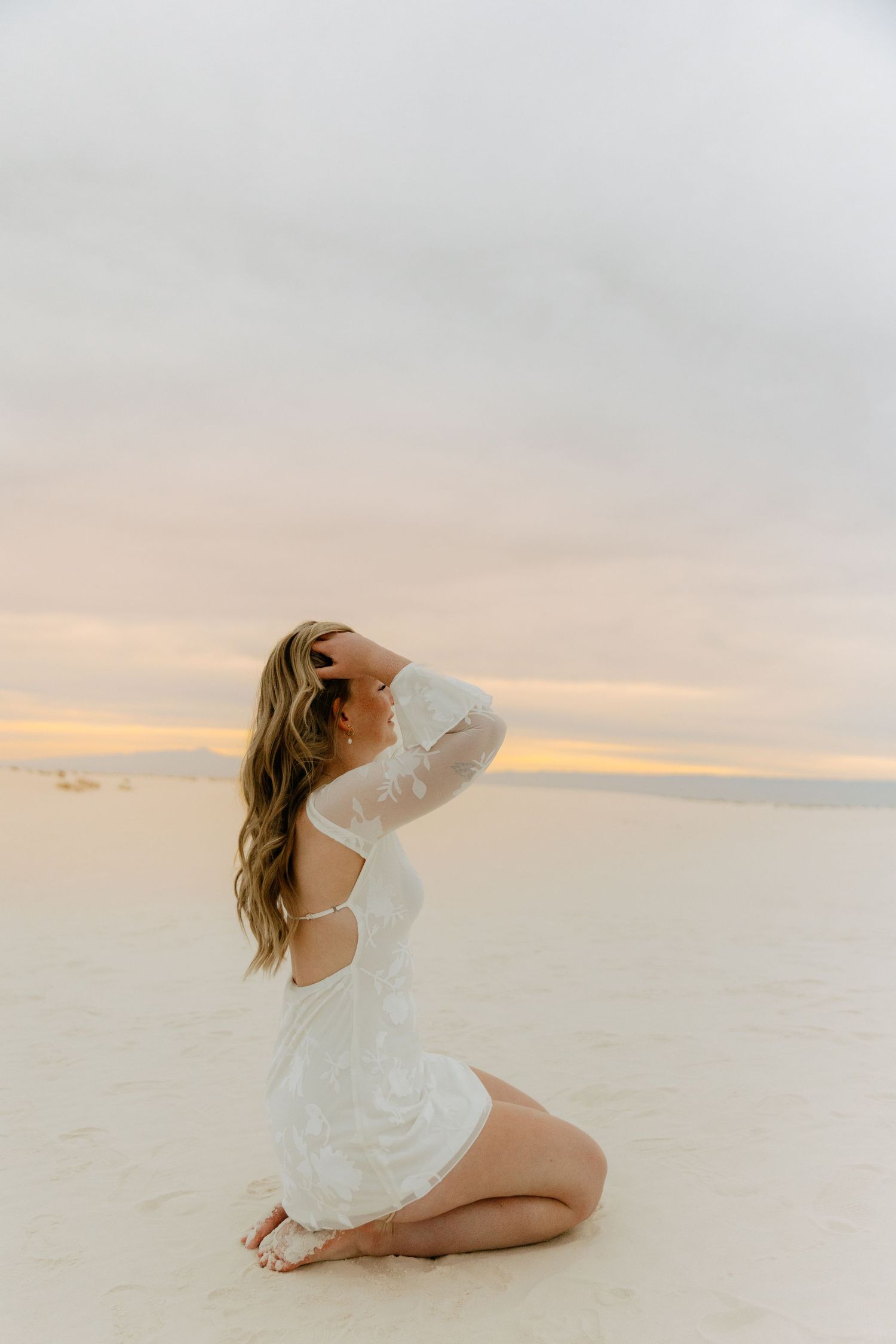 Woman in white lace mini dress kneels on white sand dunes with hands in hair at sunset.