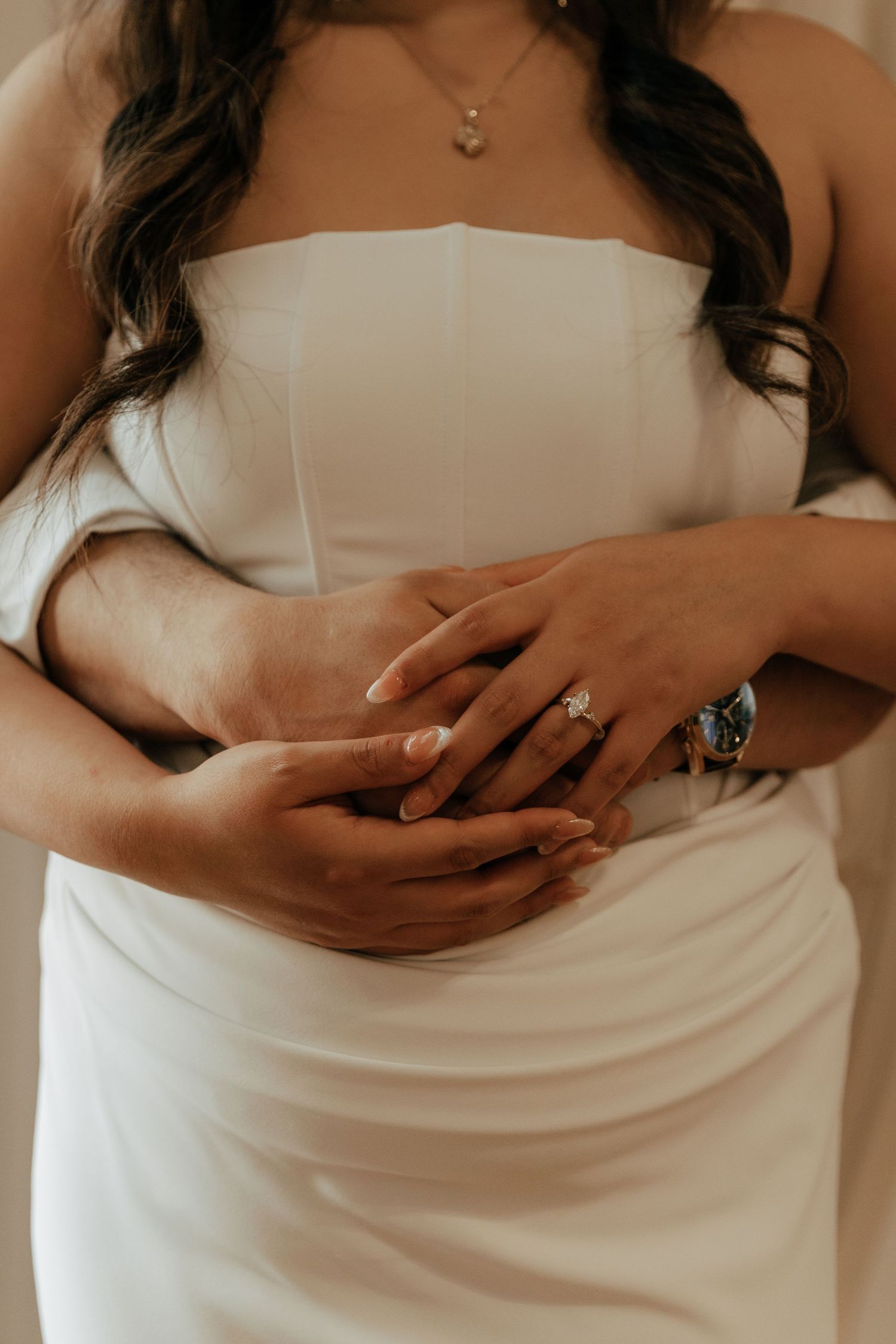 Bride in white strapless gown with groom's arms wrapped around her, showcasing diamond engagement ring and watch.