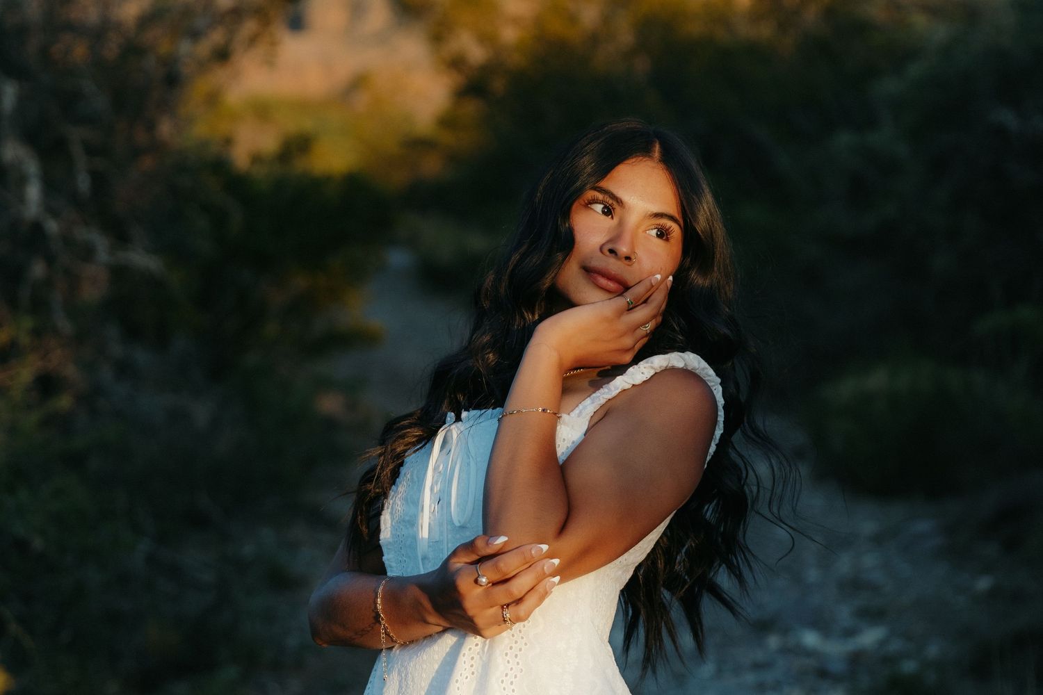 Young woman in white dress posing thoughtfully in golden hour sunlight among lush greenery.