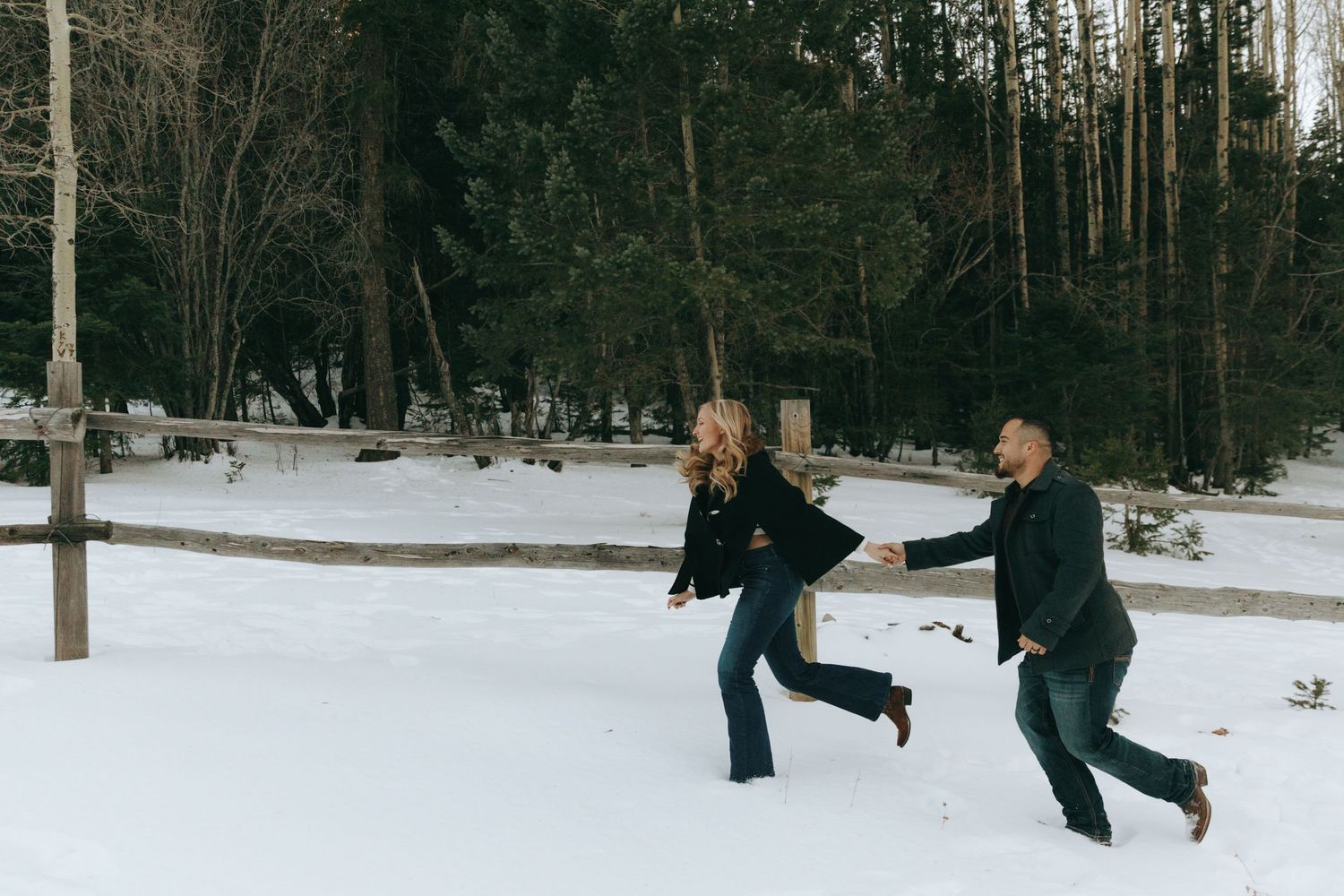 Couple holding hands and running playfully through snow in a winter forest during an engagement photo session.