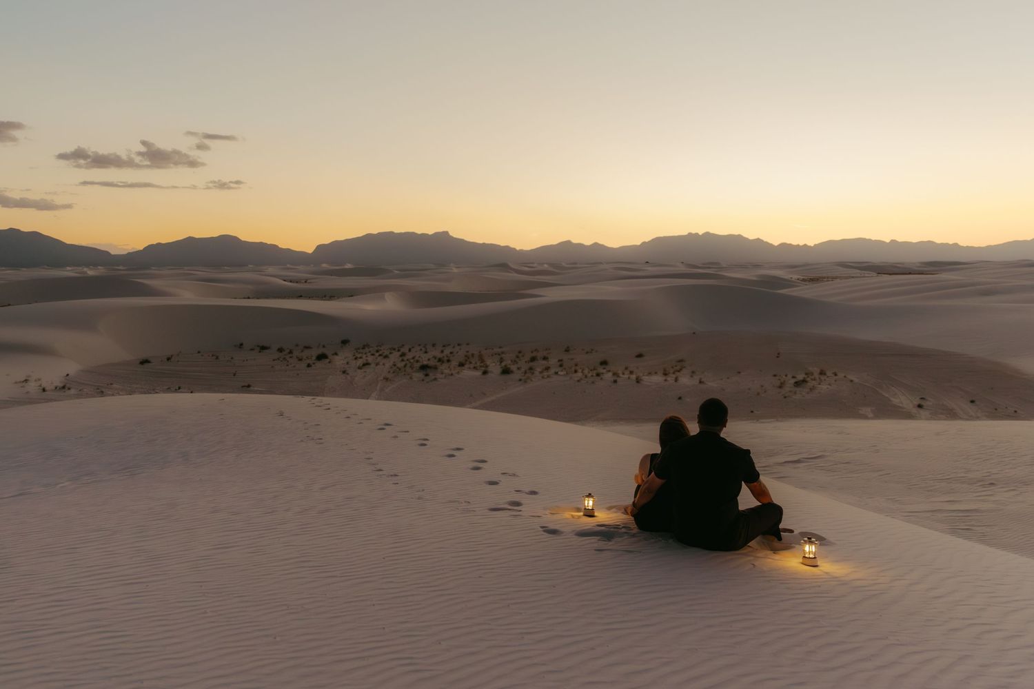 Couple sitting together on white sand dunes at sunset with candles, enjoying a romantic desert landscape view.