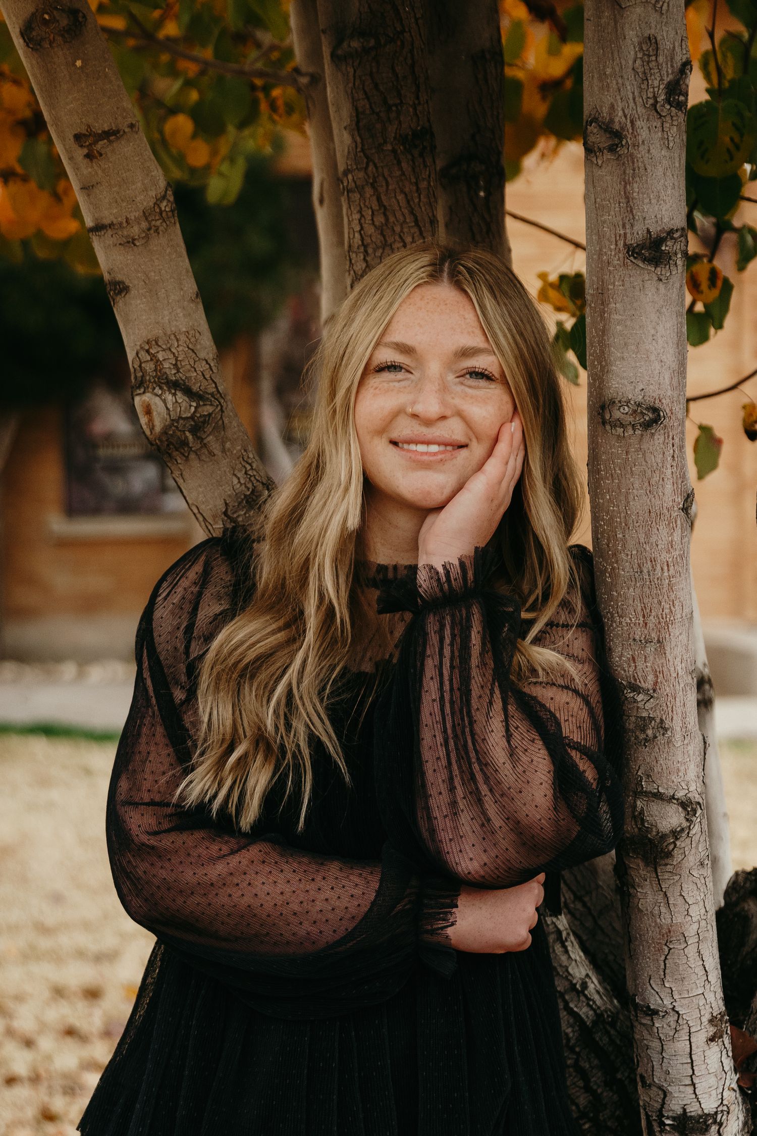 Blonde woman in black sheer dress leaning against birch trees smiling in autumn setting.