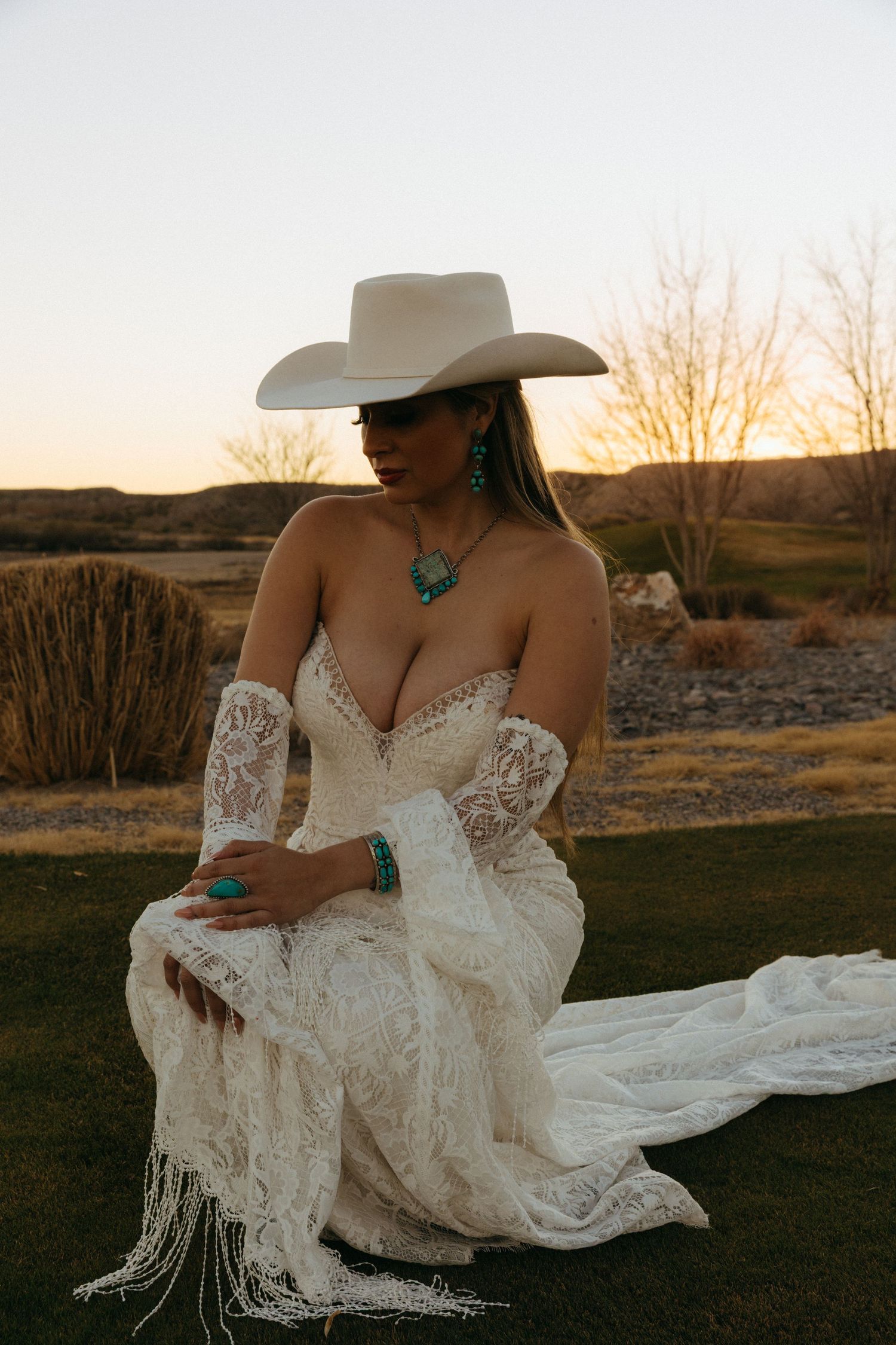 Bride in strapless lace gown with turquoise jewelry and white cowboy hat at golden hour.