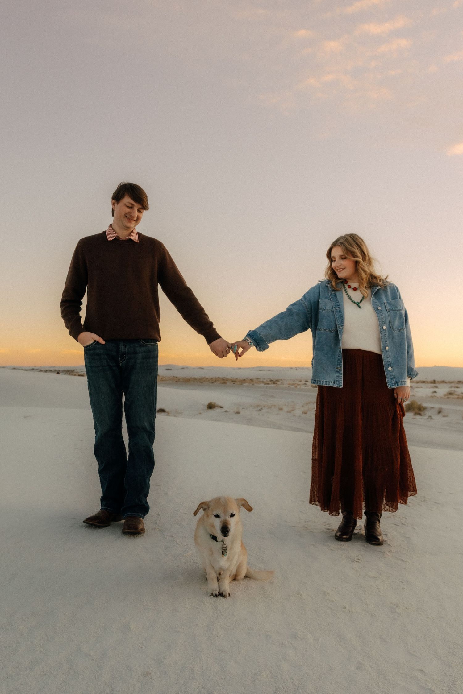 Young couple holding hands with their dog on white sand dunes during a colorful sunset in the desert.