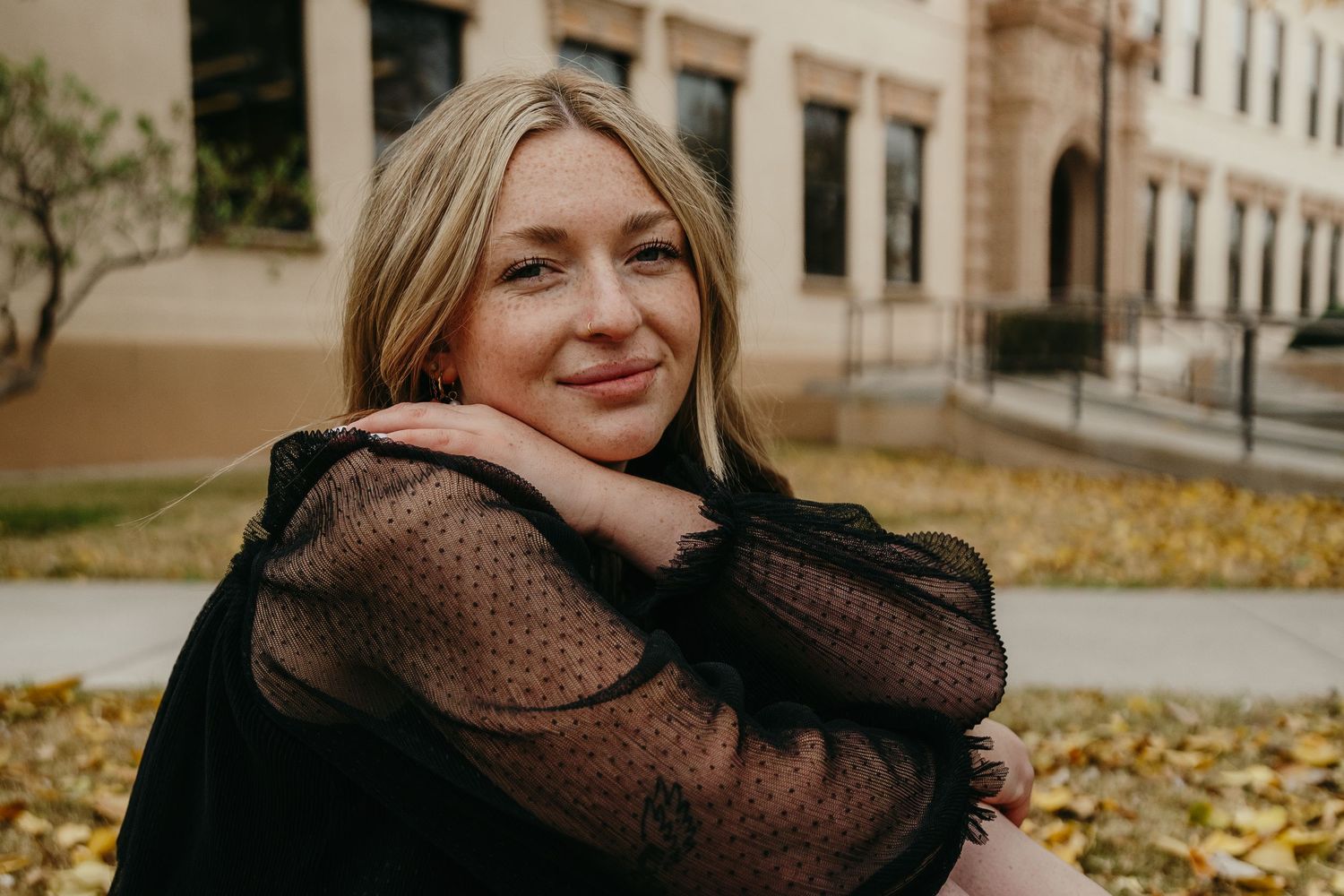 Smiling blonde woman in black sheer top sitting outdoors surrounded by fallen leaves.