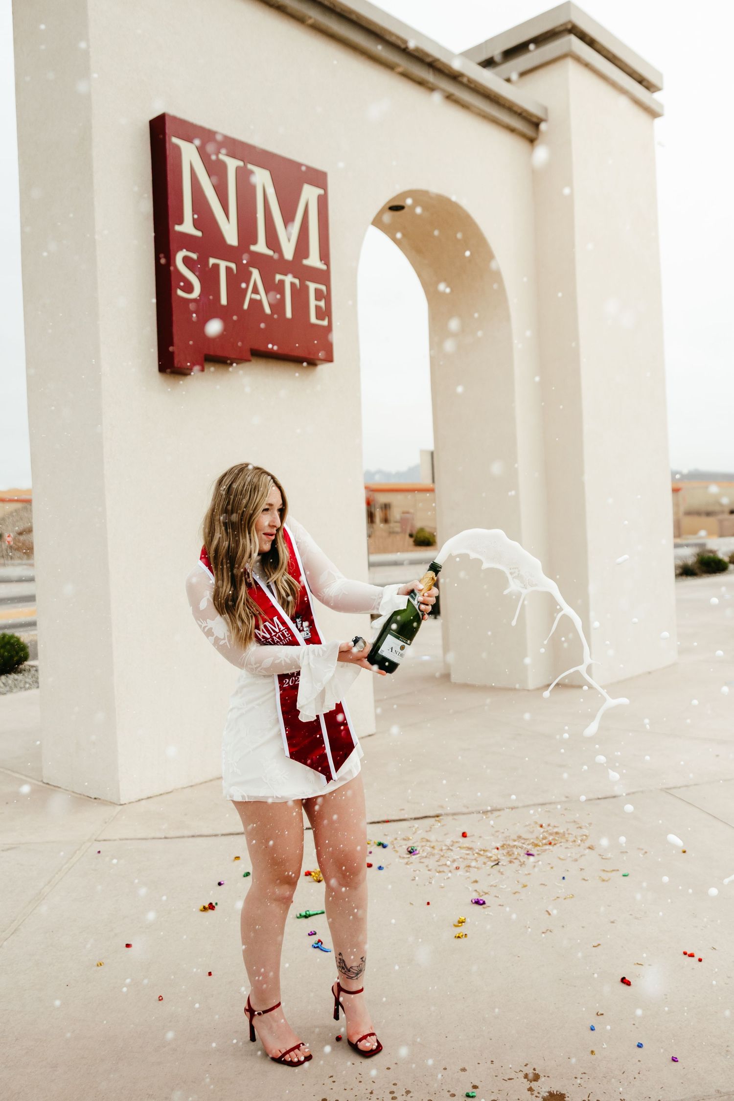 Graduate in white lace dress popping a champagne bottle with confetti in front of NM State University sign.