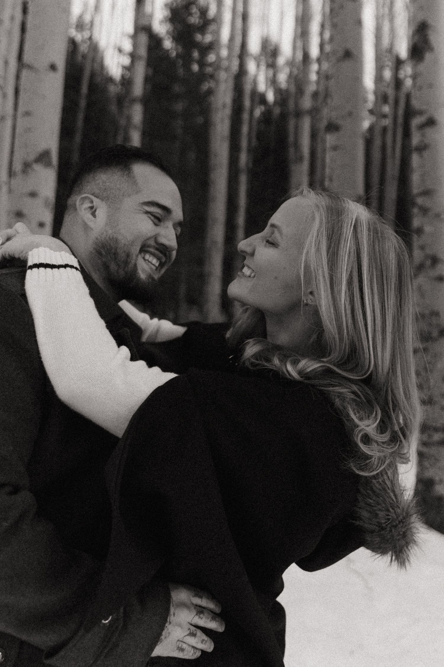 Black and white photo of a laughing couple embracing in a snowy aspen forest during winter.