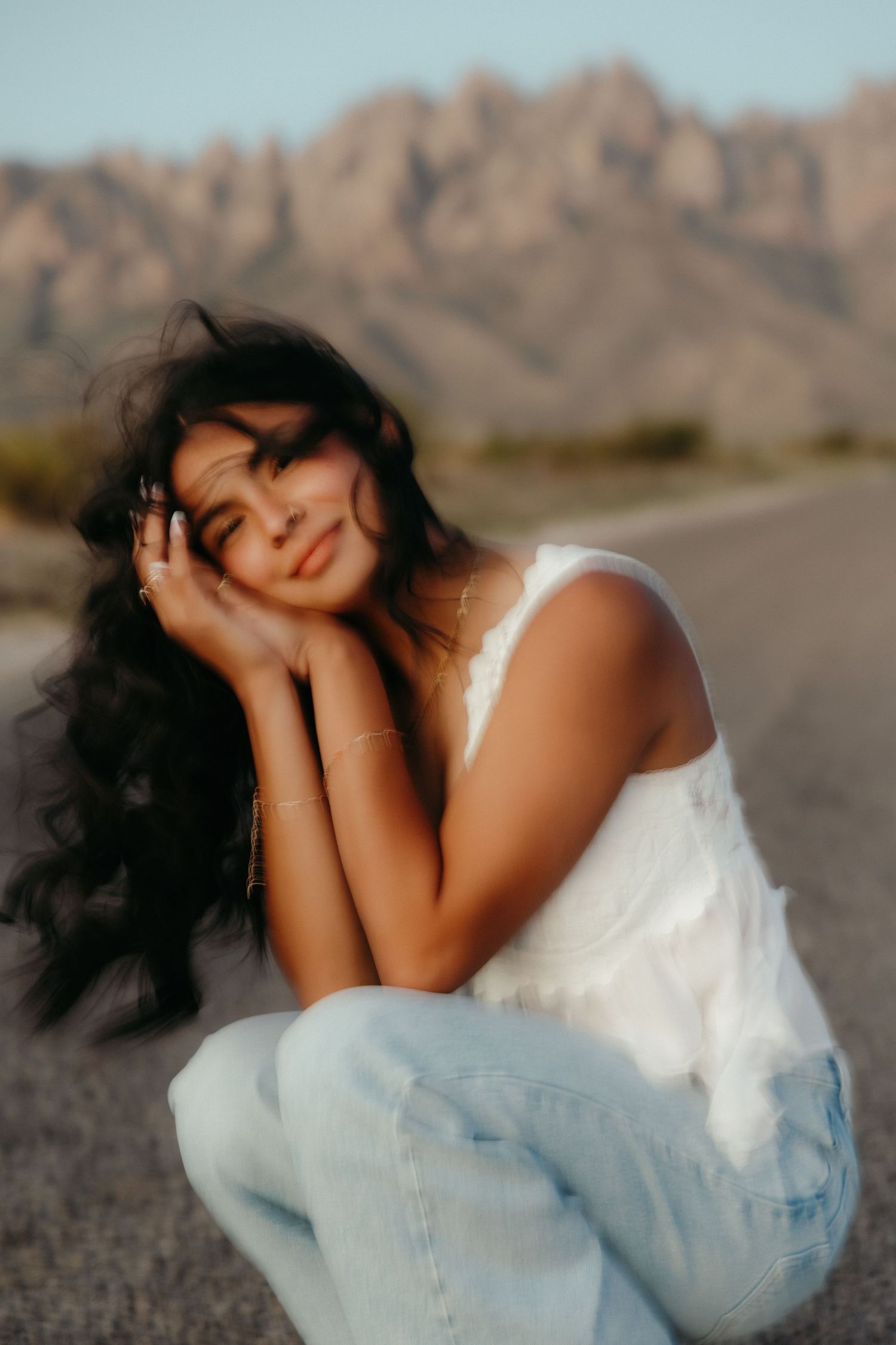Young woman in white tank and jeans poses on desert road with mountain backdrop at golden hour.