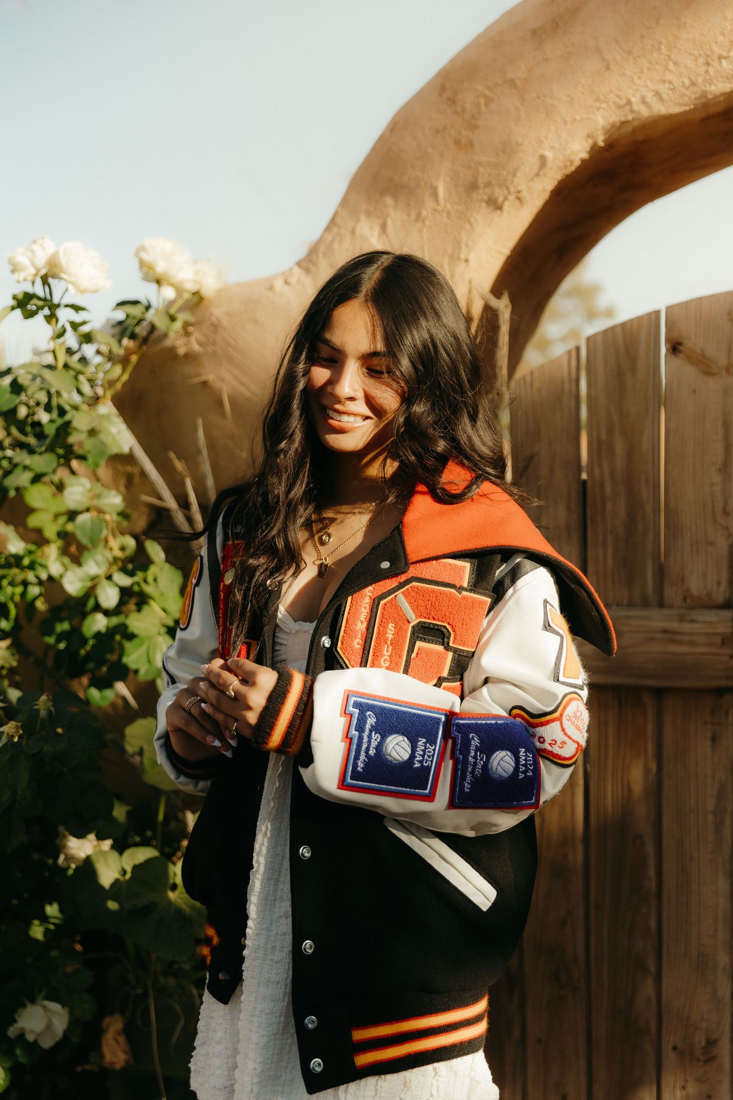 Young woman smiling in orange varsity jacket holding graduation items outdoors near roses and adobe arch.