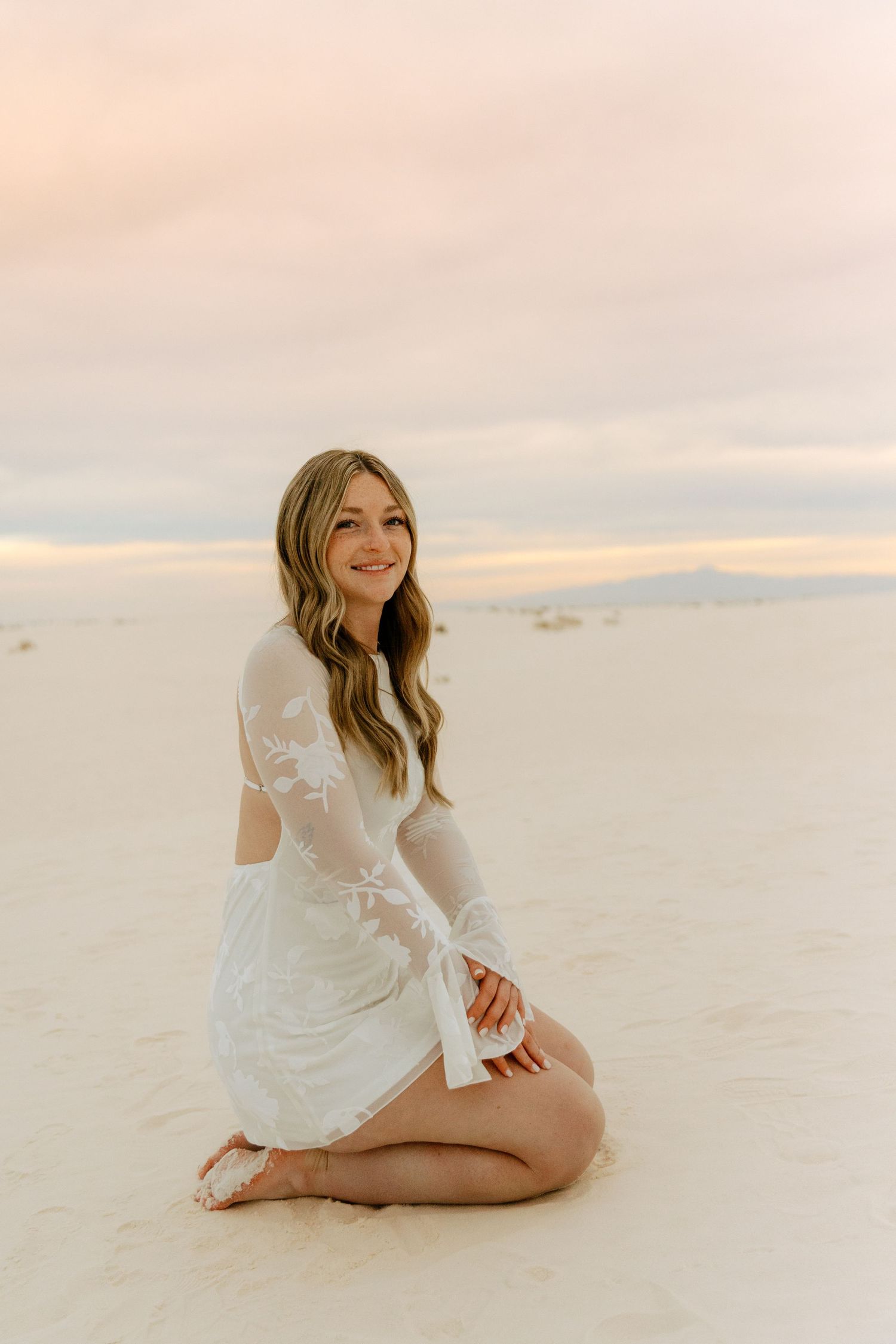 Smiling blonde woman in white lace dress sits gracefully on white sand dunes at golden sunset.